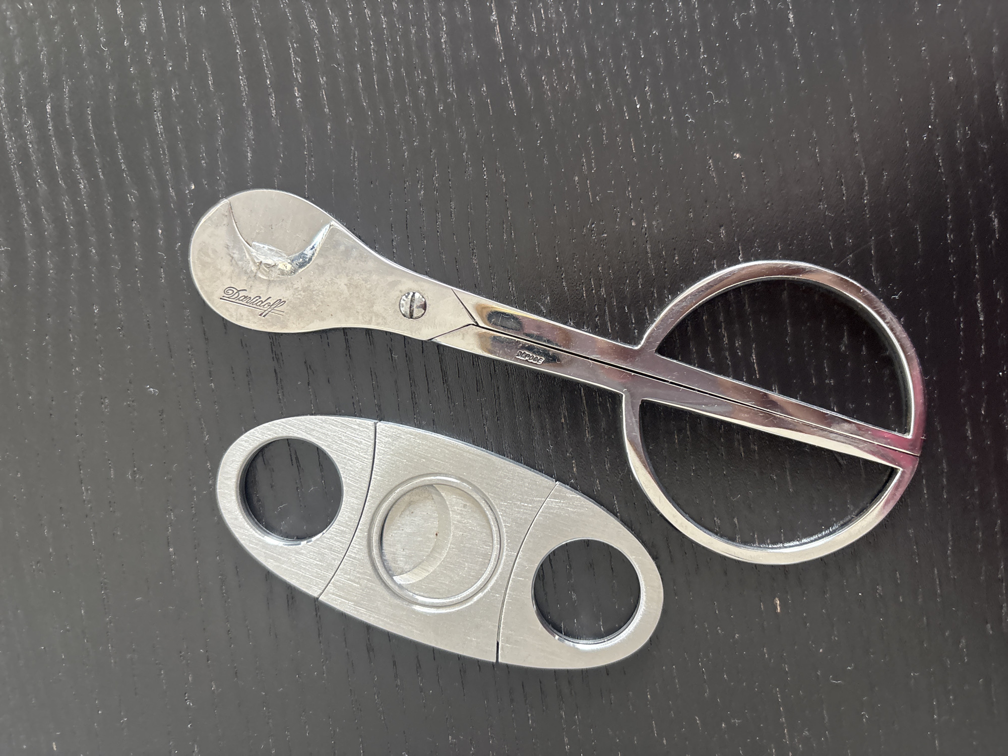 The image displays a top-down view of two silver cigar accessories on a dark, textured surface, likely a wooden table. On the left is a silver guillotine-style cigar cutter with two finger holes and a circular cutting mechanism. To the right is a pair of silver cigar scissors, also with finger holes. The scissors are open, and the blades are visible. The top handle of the scissors has an engraved logo that appears to say "Davidoff" and a small image of a bird. The stem of the scissors has a small screw and is also etched with text, possibly a model number, that reads "2F-005". The background surface has visible wood grain and some dust particles. The scene is well-lit, with a slight reflection on the metallic objects. There is no indication of the city of Dubai, United Arab Emirates, in the image.