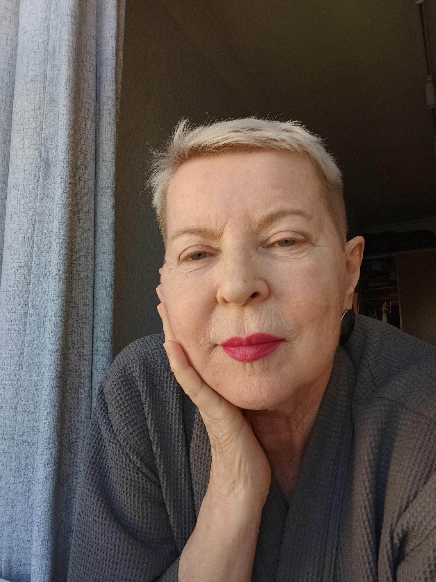 A close-up, head-and-shoulders selfie features an older woman with short, blond hair, wearing a gray waffle-knit robe and bright pink lipstick. Her left hand is propped under her chin, and she looks directly at the camera. A gray curtain hangs on the left side of the frame, and the background is dark, with some items visible in what appears to be a closet on the right. The lighting suggests natural light coming from the left. The location in Saint Petersburg, Russia is not discernible from the image.