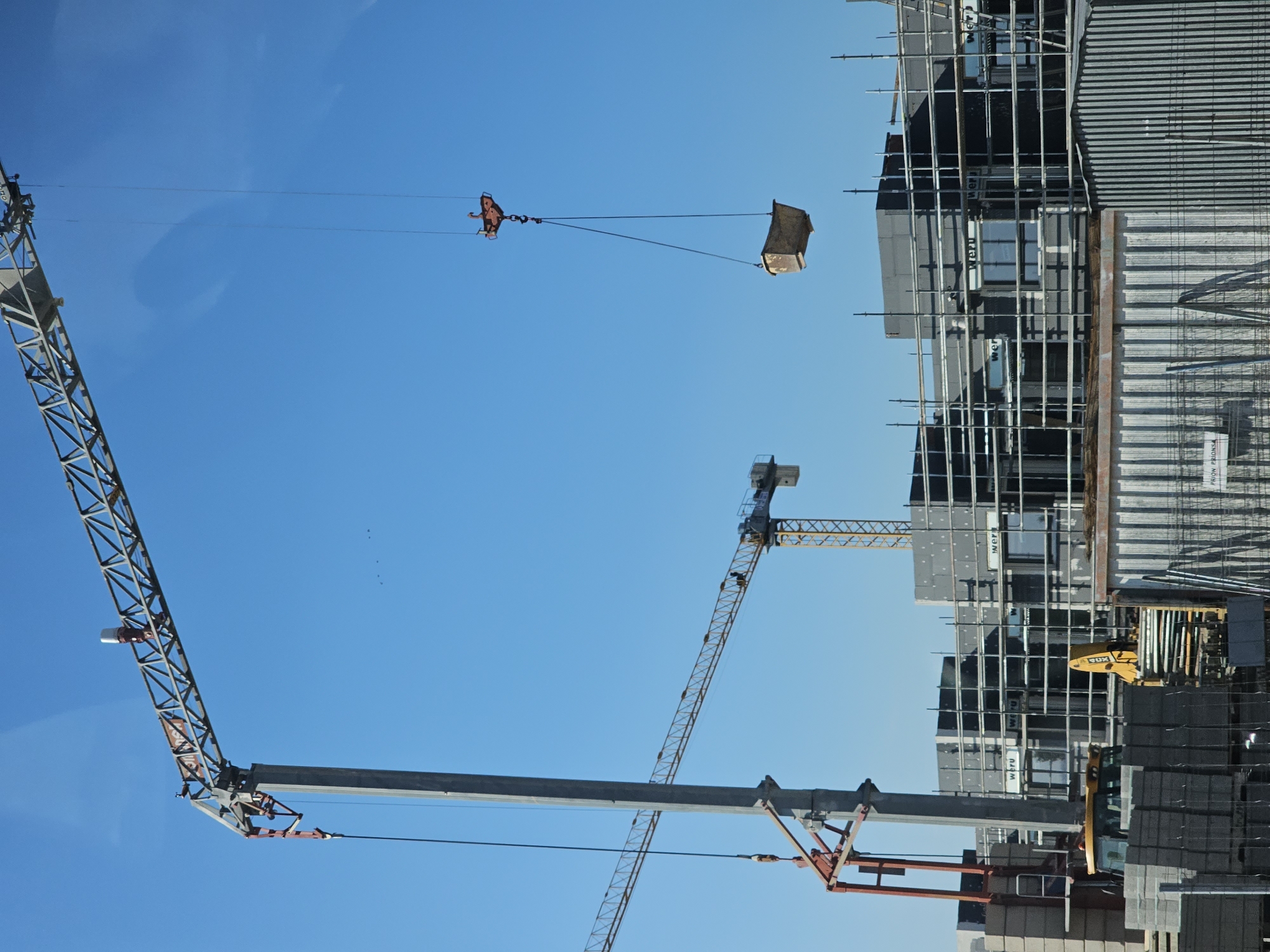 This image depicts a construction site in Cuarte de Huerva, Spain, under a clear blue sky. Two tower cranes are prominently featured. The larger crane on the left has a significant boom extending upwards and to the right. A smaller, red-colored attachment is visible near the base of its vertical support. A concrete box is suspended in the air from the larger crane by cables. In the middle ground, a yellow construction vehicle, possibly a backhoe loader, is partially visible. The construction site itself shows building structures in various stages of completion, characterized by dark grey exterior panels and scaffolding. Several windows with white frames are present on the buildings, some bearing the "WERU" logo. In the foreground, stacks of building materials, including what appear to be concrete blocks and thin planks, are organized. A corrugated metal structure is situated in the lower right portion of the frame. The overall scene suggests active construction work.