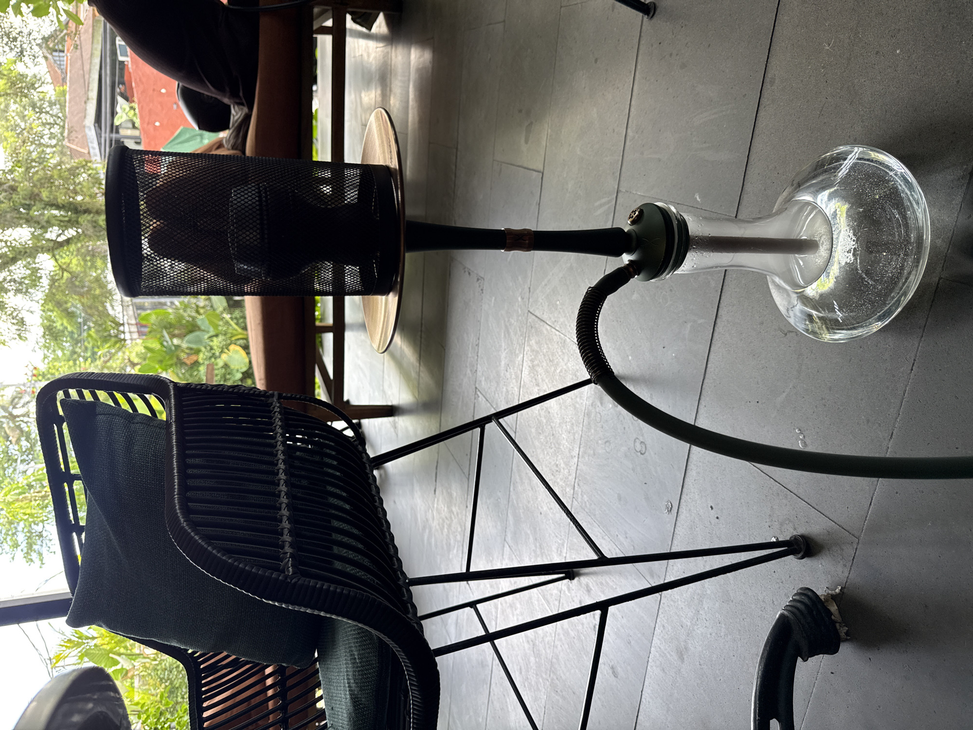 The image depicts an outdoor setting, likely a patio or terrace, in Ubud District, Indonesia. The primary focus is on a hookah (water pipe) placed on the gray tiled floor. The hookah consists of a glass base filled with water, a central metal stem with a decorative accent, a tray, and a mesh-covered bowl. A black hose extends from the stem. Beside the hookah, there's a black rattan chair with a dark cushion. A person is partially visible in the background, seated at a wooden table. The surrounding environment includes lush greenery, partially visible through an opening, suggesting a natural setting. The buildings appear to be residential, with their architectural style hinting at the regional aesthetic. The overall setting suggests a relaxed or recreational space.