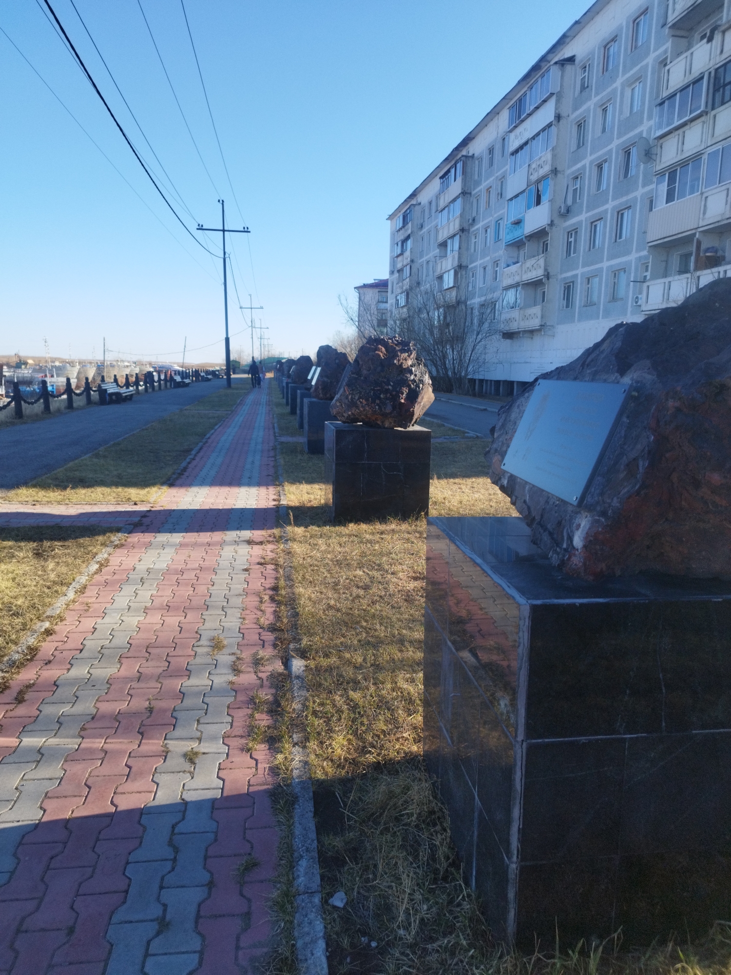 The image shows a view of a section of the embankment in the urban-type settlement of Zhatay, Sakha Republic, Russia. A pedestrian walkway paved with red and grey paving stones stretches into the distance, lined on one side with irregularly shaped dark rocks mounted on dark, polished, rectangular pedestals. Many of the stones have metal plaques affixed to them. Patches of dry grass flank the walkway and pedestals. Beyond the stones and grass is a paved area bordering the riverbank, with a dark fence, benches, and what appear to be boats docked in the background. On the opposite side of the walkway from the river is a large apartment building with light-colored walls and multiple windows and balconies. Overhead are utility poles and lines against a clear, bright blue sky. In the background, there are people walking along the embankment.