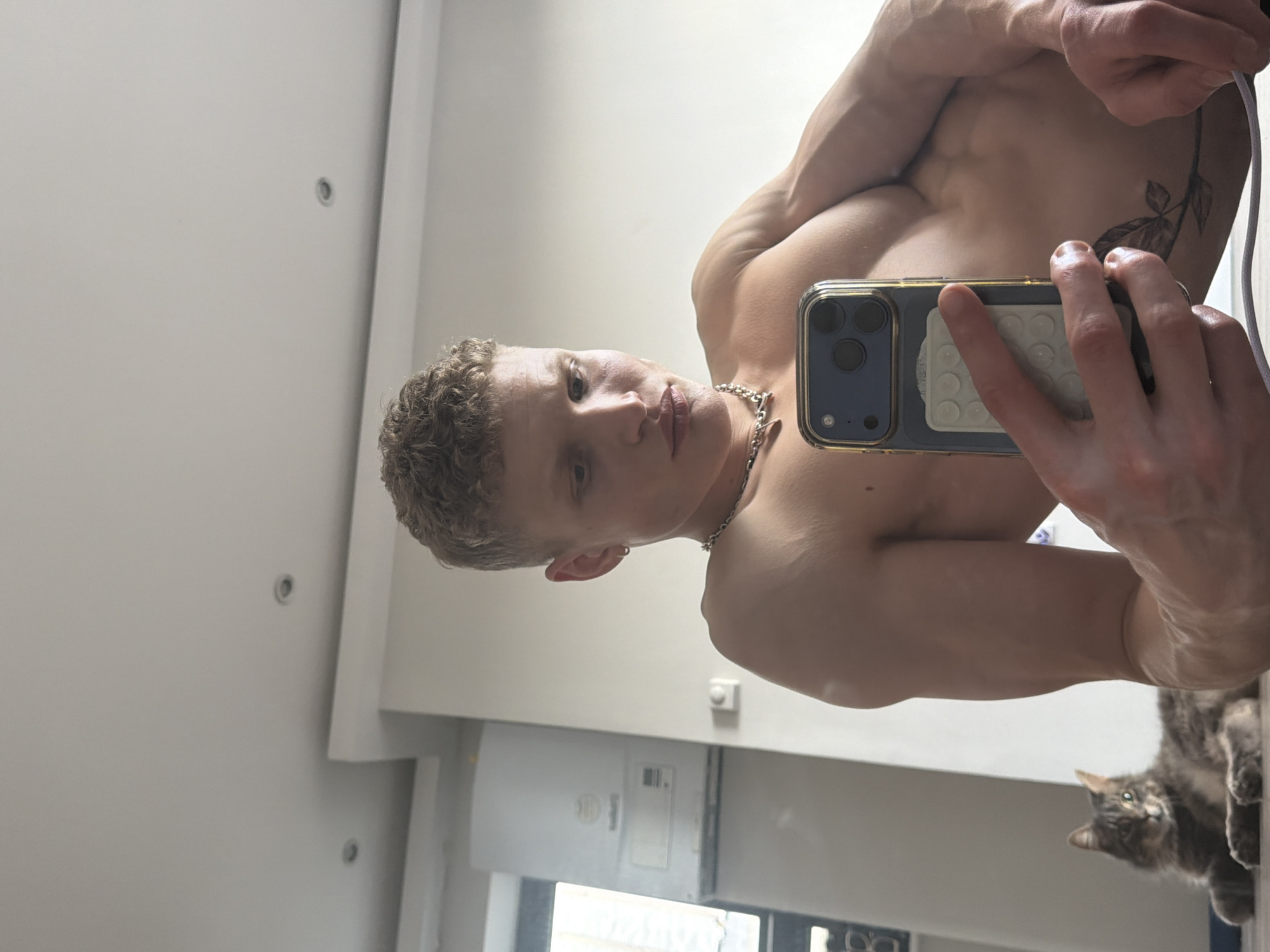 A shirtless man with short curly hair and a silver chain is taking a selfie in a room with white walls. He is holding an iPhone in his right hand. A cat is visible in the lower right corner of the image. A window is visible in the background. The location cannot be determined from the image, only that it is likely indoors, in a room with white walls.