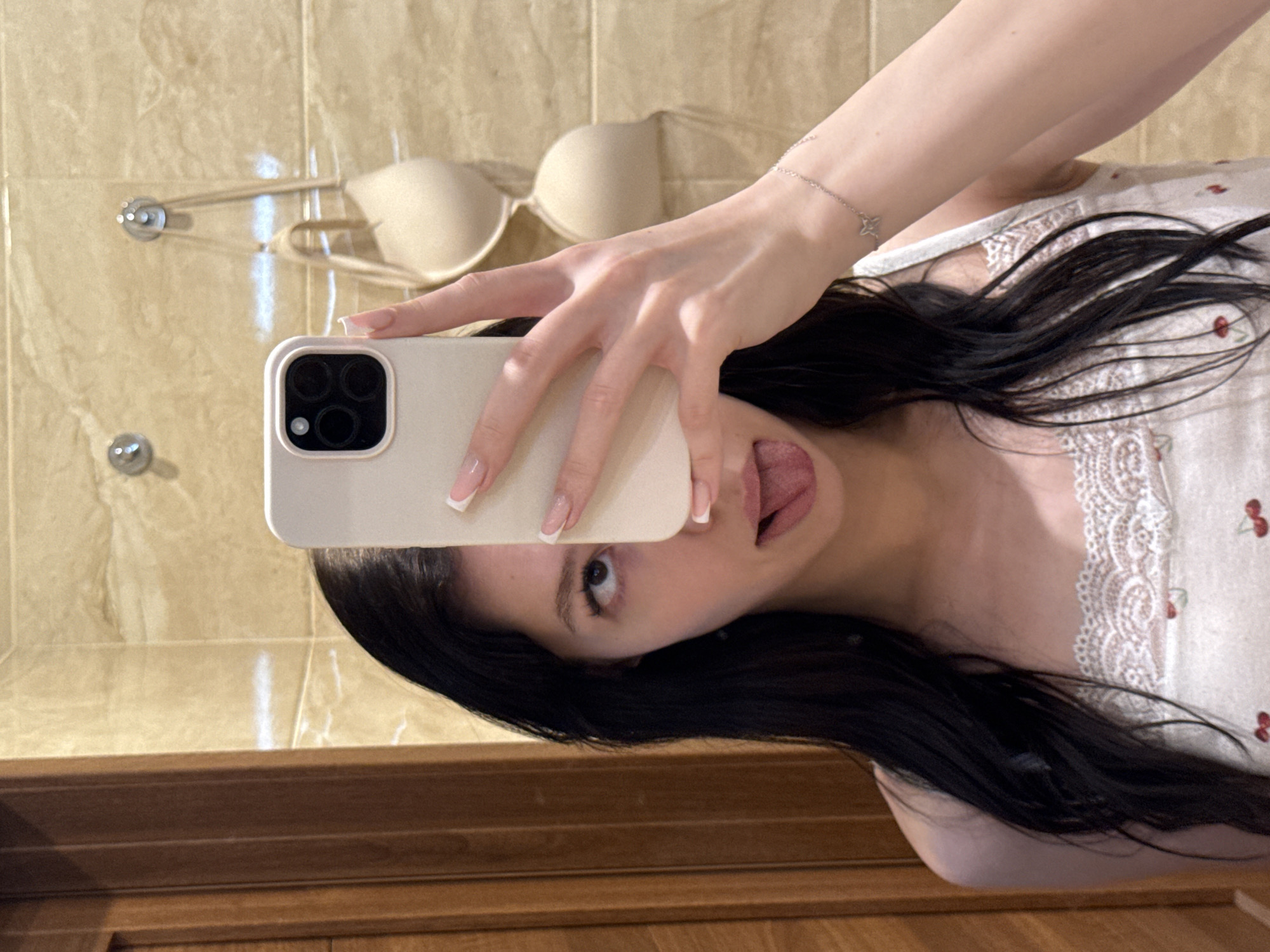 A young woman with dark hair is taking a selfie in what appears to be a bathroom. She is holding a phone in her right hand, covering the upper half of her face. Her tongue is sticking out, and her left eye is looking upwards. She is wearing a white tank top with a lace trim and a cherry pattern. In the background, on a tiled wall, a beige bra is hanging from a hook. The overall environment suggests a private domestic setting, likely a bathroom. There is no information to suggest this location is in Khimki, Russia.