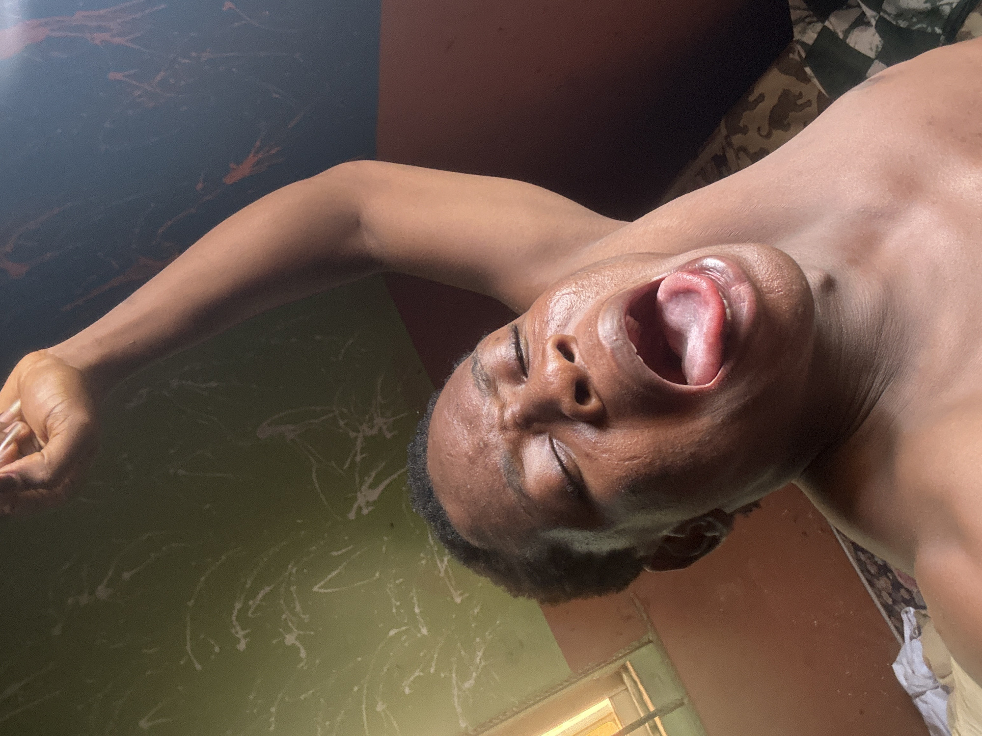 An individual with dark skin is captured in a close-up, diagonal shot, seemingly mid-yawn or stretch. Their eyes are closed, mouth wide open revealing the tongue and inner mouth, conveying a sense of deep relaxation, fatigue, or boredom. One arm is extended upwards and to the left, with the hand partially visible, suggesting a stretch. The person appears shirtless, exposing their upper torso and shoulder.
The setting is indoors, likely a room with varying wall colors and textures. In the bottom left, a light green wall is visible, adorned with irregular white splatters or abstract paint marks. Towards the top left, a darker, possibly black or dark brown wall with faint reddish markings is present. On the right side, behind the person's head and shoulder, a patterned fabric or surface is seen, featuring both a black and white checkered design and a camouflage-like print. A window, admitting warm light, is visible in the bottom right corner, suggesting daytime. The lighting in the room is somewhat dim overall, with direct light highlighting parts of the person's face and body. No text is visible in the image.