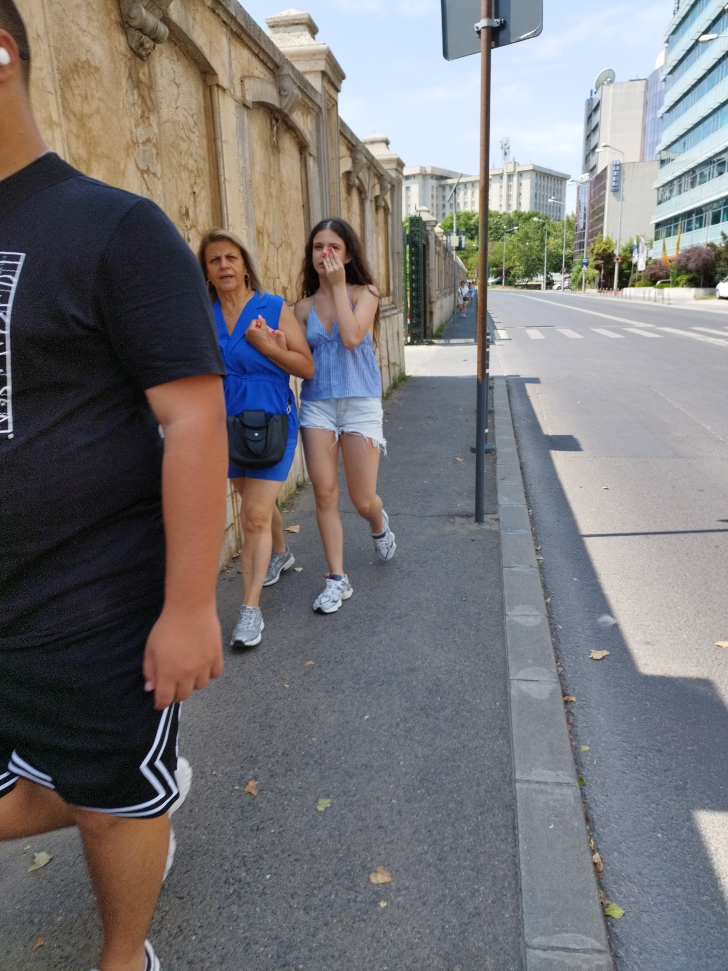 A medium shot captured from a low angle shows two women walking on a sidewalk in front of an ornate, textured wall. The woman on the left, with blonde hair and a blue dress, holds her hand near her face. She wears a black purse crossbody. The woman on the right, with dark hair, wears a blue and white striped tank top and denim shorts. She is covering her mouth with her hand. A person in the foreground, out of focus, wears a black t-shirt and shorts. They are walking away from the camera. The sidewalk is paved with grey concrete, and a street runs alongside it. In the background, modern buildings with glass facades and older, utilitarian buildings are visible. Trees line the street. The sky is clear and blue. The image appears to be taken on a sunny day in an urban setting, likely Bucharest, Romania, given the architectural style and urban landscape.