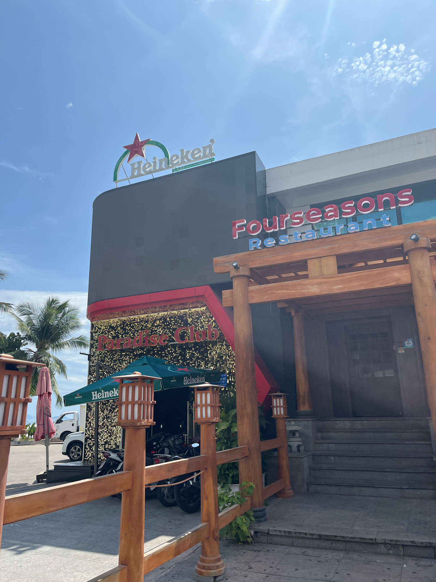 The image shows the facade of a building with two businesses: "Fourseasons Restaurant" and "Paradise Club." The "Fourseasons Restaurant" sign is large and red, with "Restaurant" written in blue underneath. The building has a Japanese-inspired entrance with wooden pillars and stairs. To the left, "Paradise Club" is advertised with a glittering gold background and red lettering. A Heineken logo is visible on a green awning covering an outdoor seating area. Several motorcycles are parked under the awning. Palm trees are visible in the background, suggesting a tropical location. The sky is blue with some scattered clouds. The location is likely along a street in Nha Trang, Vietnam, given the signage and tropical vegetation.