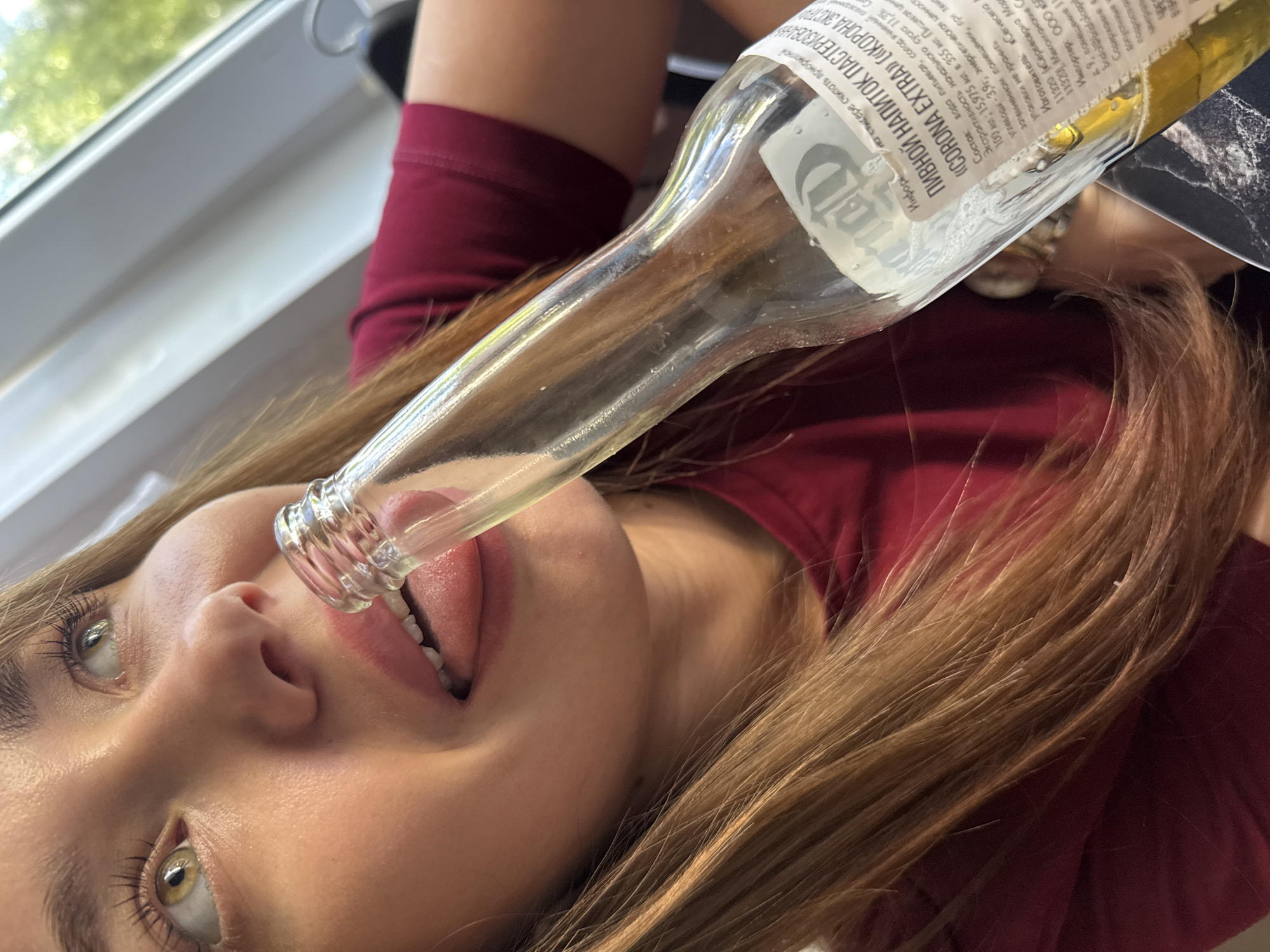 This media file is a close-up, vertically oriented shot of a young woman. She has fair skin, light brown hair, and light-colored eyes. The woman is wearing a dark red top. Her mouth is open, and her tongue is visible, interacting with the narrow neck of a clear glass bottle, which is angled towards her face from above. The bottle has a label with visible text in Russian, including "«CORONA EXTRA»" and ingredient information like "Пищевая цен" (nutritional value) and "углеводы 3,9г" (carbohydrates 3.9g). The background is blurred, but appears to be indoors with a window or light source showing some greenery outside. The overall impression is a playful or experimental interaction with the bottle. The visible text on the bottle indicates the product is "Corona Extra" and lists manufacturing and import details, suggesting a retail or consumption setting, possibly in Chaikovsky, Russia, based on the provided location context. The lighting suggests it could be daytime.