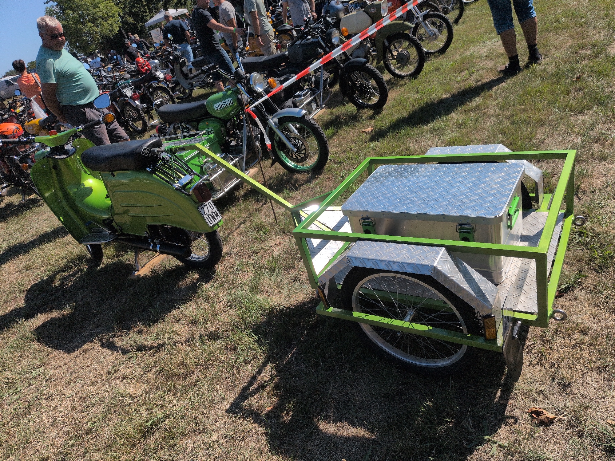 The image shows a gathering of people and vintage motorcycles in Hohenferchesar, Germany. In the foreground, a bright green scooter is parked next to a green trailer equipped with a silver toolbox. The scooter has a license plate that reads "270 UMW". Behind the scooter, several other motorcycles are visible, along with a crowd of people milling around. The event appears to be an outdoor festival or rally. The ground is covered in dry grass, suggesting a sunny and warm day.