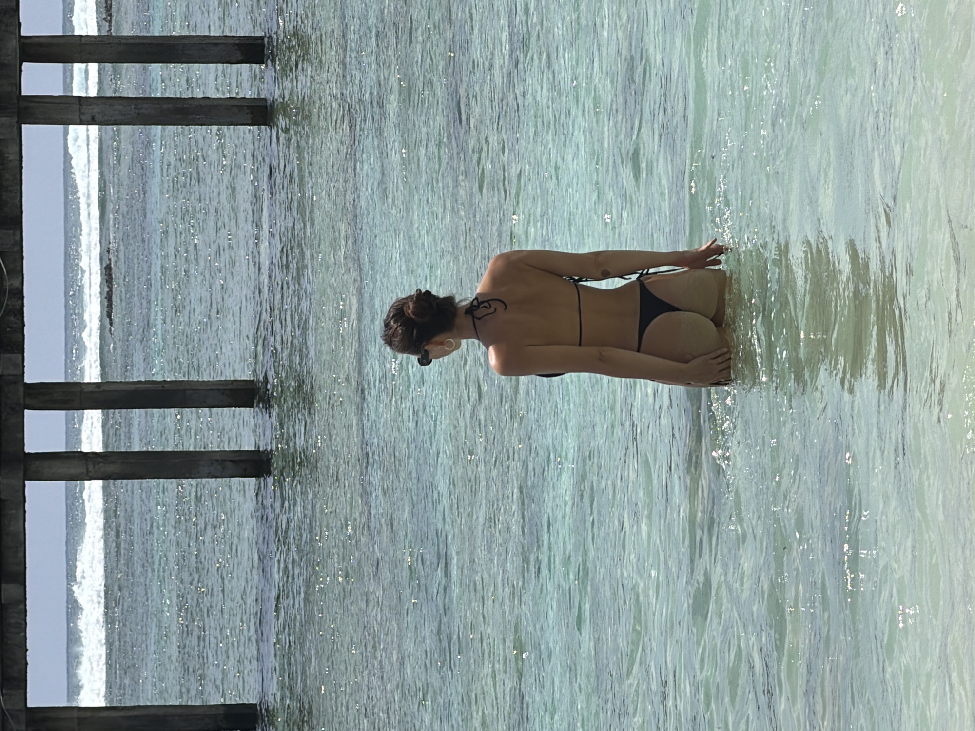 The image captures a woman from behind, kneeling or sitting in shallow, clear, turquoise water. She is positioned slightly to the right of the frame's center, facing away from the camera. Her hair is tied back in a bun, and she is wearing a black string bikini with her hands resting in front of her. Small hoop earrings are visible.
The water is sparkling with sunlight reflections, showing gentle ripples and hints of the sandy seabed beneath. To the left of the frame, several dark, rectangular pillars, likely part of a pier or similar wooden structure, extend vertically into the water. Beyond these pillars and further out, white-capped waves are visible, suggesting an open ocean or sea.
The scene depicts a sunny daytime environment, indicating warm weather. The overall impression is one of relaxation at a tropical coastal location. There is no visible text in the image. Given the context, this scene is likely in General Luna, Philippines.