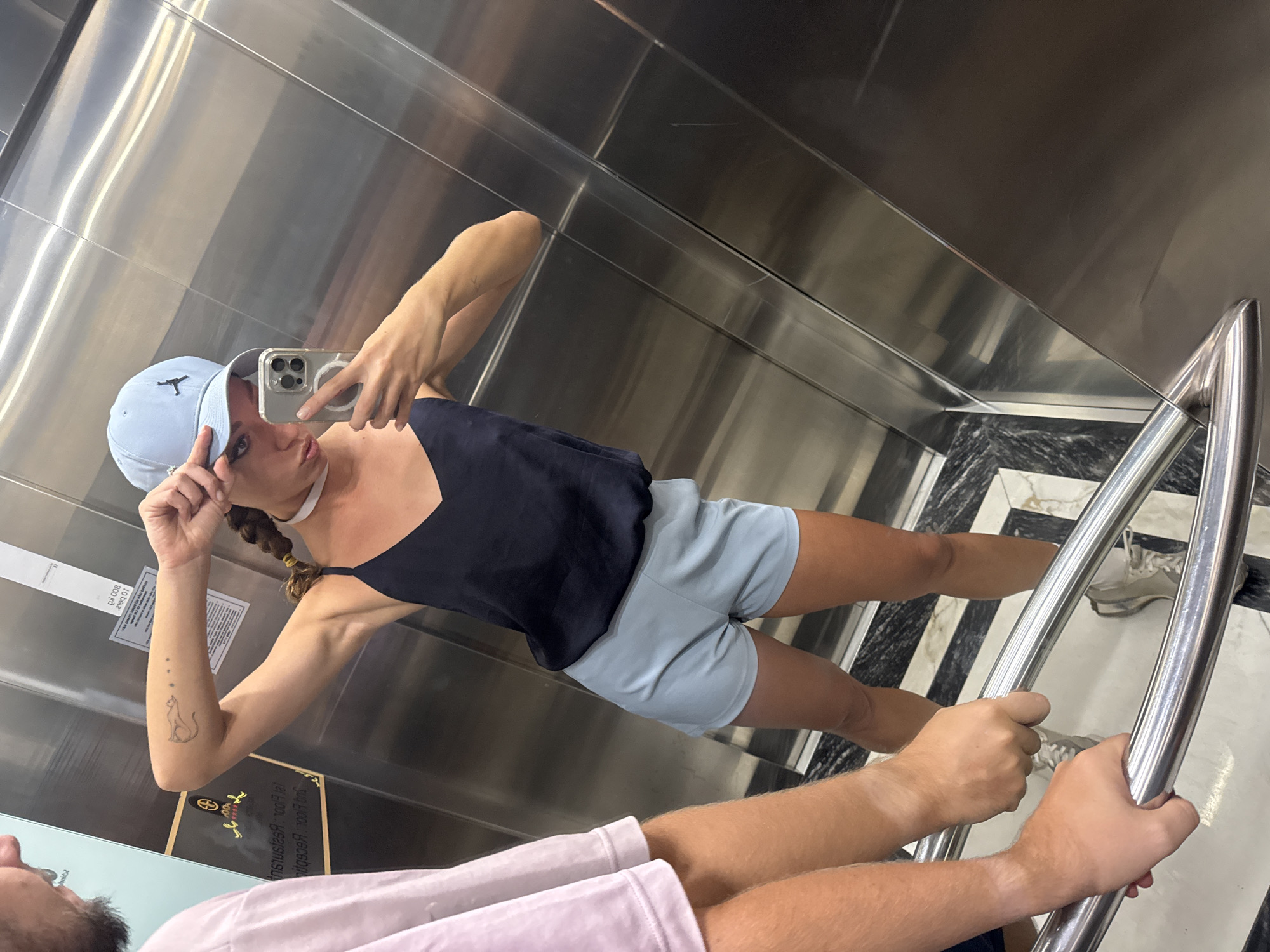 A medium shot taken inside an elevator reveals a woman taking a selfie. She is wearing a light blue baseball cap, a navy tank top, and light blue shorts. She is holding a smartphone, presumably taking the picture. Her left arm is raised, showing a tattoo of a cat on her forearm. She has light skin, and her hair is braided. In the lower portion of the image, a man is reflected in the mirrored wall of the elevator. He is wearing a light pink t-shirt and is holding onto the elevator's handrail. The elevator interior is comprised of brushed metal walls and a black and white patterned floor. It is not possible to confirm the location as Hà Nội, Vietnam from the visual information.