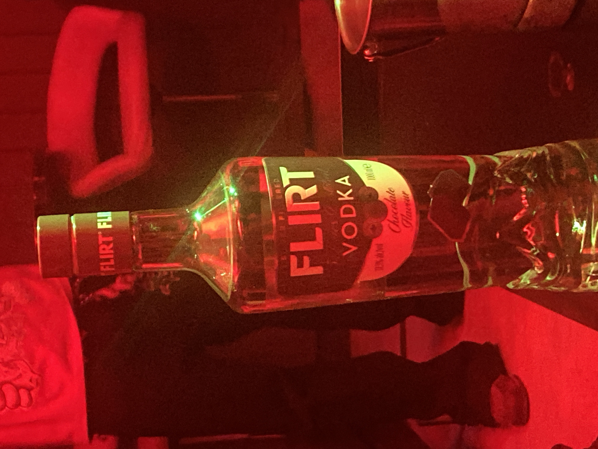 A close-up, eye-level shot features a clear glass bottle of Flirt Vodka with a black label. The label prominently displays the brand name "FLIRT" in large, white, block letters, with "VODKA" written below it in a smaller, white font. The bottle is illuminated from within by green lights, which wrap around its neck and upper body, creating a vibrant effect against the dark background. The lower part of the label includes text that appears to indicate "Chocolate Flavour" and "1000 ml".
The bottle stands on a dark surface, possibly a table or bar. To the right of the bottle, a portion of a metallic container, perhaps an ice bucket or a shaker, is visible. In the background, blurred figures and furniture are discernible, bathed in a strong red light. The overall ambiance suggests a dimly lit, possibly club or bar setting. The red lighting casts a reddish hue over the entire scene, giving a somewhat abstract and atmospheric quality. The image is consistent with a scene that could be found in a nightlife establishment in Abuja, Nigeria, given the style of lighting and typical bar setup.