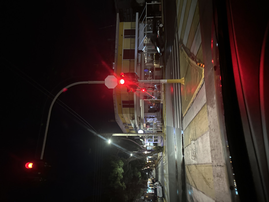 The media file presents a nighttime urban street scene, captured from a slightly elevated perspective, possibly from within a vehicle, with a partial view of a car interior frame on the right. The image, when oriented correctly, shows a street receding into the distance under a dark, clear sky.
In the mid-ground, a prominent traffic light pole with a curved arm holds a brightly glowing red traffic signal and a white octagonal sign. Further down the street on the left, another red light is visible. The road surface below is marked with a pedestrian crossing featuring distinct white and yellow stripes, appearing wet and reflective, hinting at recent rain. Red reflections from the traffic lights are cast upon the damp asphalt.
Along the right side of the street, a multi-story building with architectural features like balconies or awnings displays yellow and reddish-brown hues, illuminated by various artificial lights emanating from inside and around it. A dark car is parked near this building. Further down the street, several streetlights cast bright beams, illuminating scattered trees and a line of parked cars along the curb. No people are visible in the frame, and there is no discernible text on any signs or structures. The scene conveys a quiet, illuminated street in Iraklio, Greece, after dark.