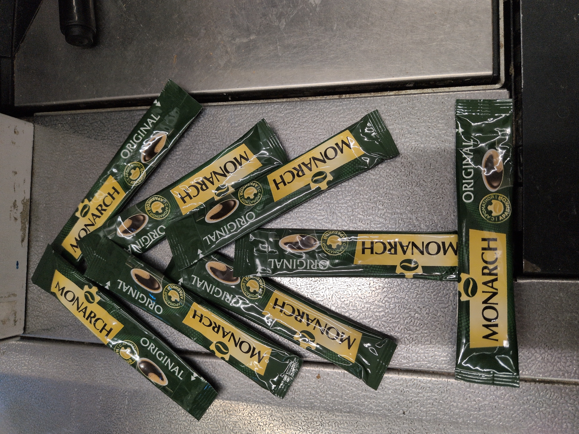 A top-down shot captures a cluster of dark green instant coffee sachets from the "MONARCH" brand, scattered across a textured, metallic surface, likely a countertop or tray, in Sochi, Russia. Each sachet prominently features the brand name "MONARCH" in yellow lettering on a horizontal yellow band. Below the brand name, the word "ORIGINAL" is printed vertically. A small circular seal, also in yellow and green, is present on each sachet, containing Cyrillic text. The most legible Cyrillic phrase, "С АРОМАТНОЙ АРАБИКОЙ," translates to "with aromatic Arabica," indicating the coffee's flavor profile. Some sachets are partially overlapping, suggesting they were loosely placed or spilled. The background reveals darker, possibly darker metallic or black edges of what appears to be part of a machine or other fixture. No people are visible, and there is no discernible activity other than the arrangement of the coffee packets. The lighting is even, suggesting an indoor setting without specific time-of-day cues.