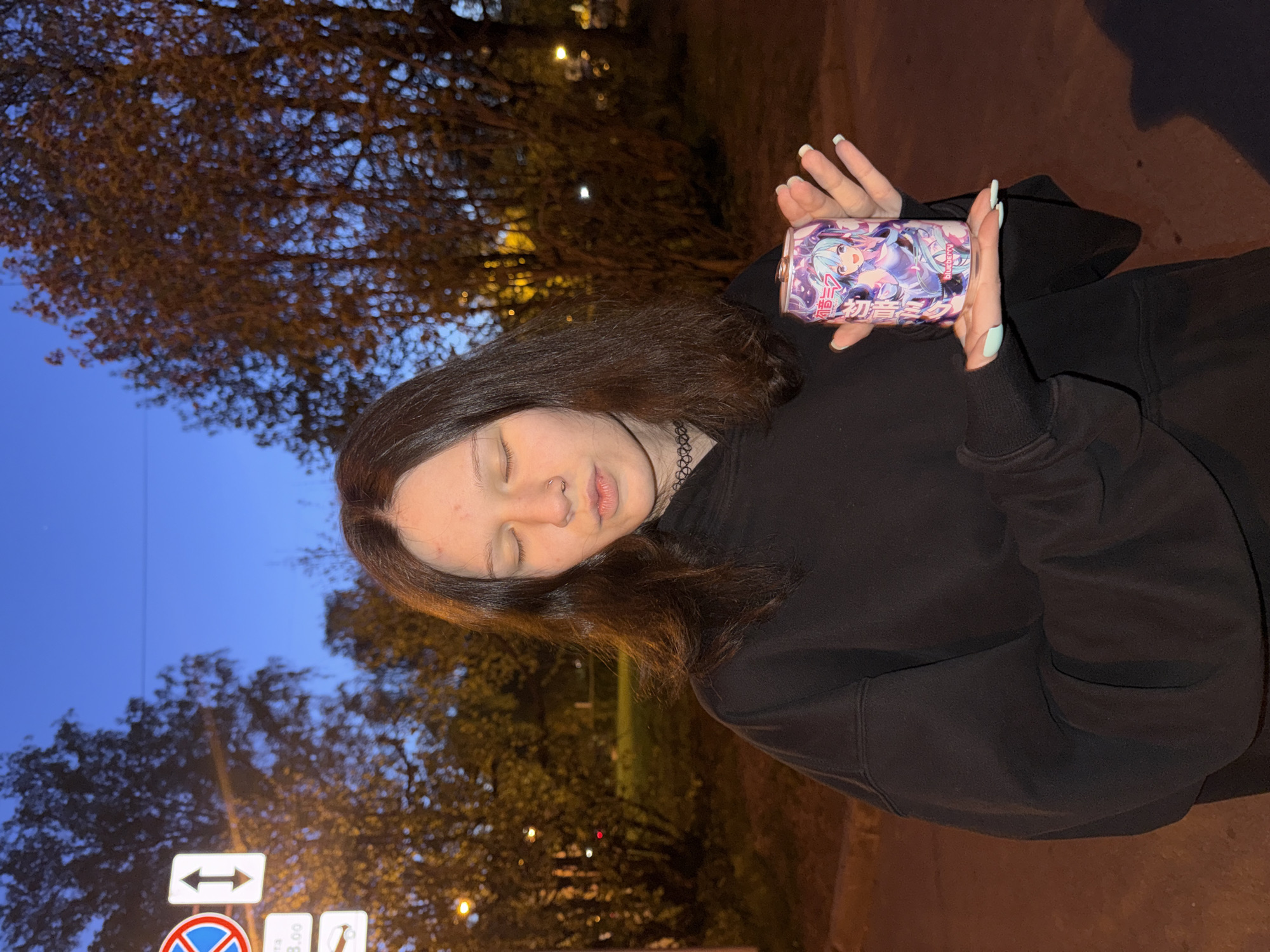 A woman is shown from the shoulders up, holding a beverage can in her left hand. She has dark brown hair, closed eyes, and a nose ring, and she is wearing a black hooded sweatshirt and a choker. She is standing on a sidewalk. The backdrop features trees and a sky gradient transitioning from dark blue to yellow, suggesting dusk. A street sign can be seen to the left of the image. The photo was taken in the city of Saint Petersburg, Russia.