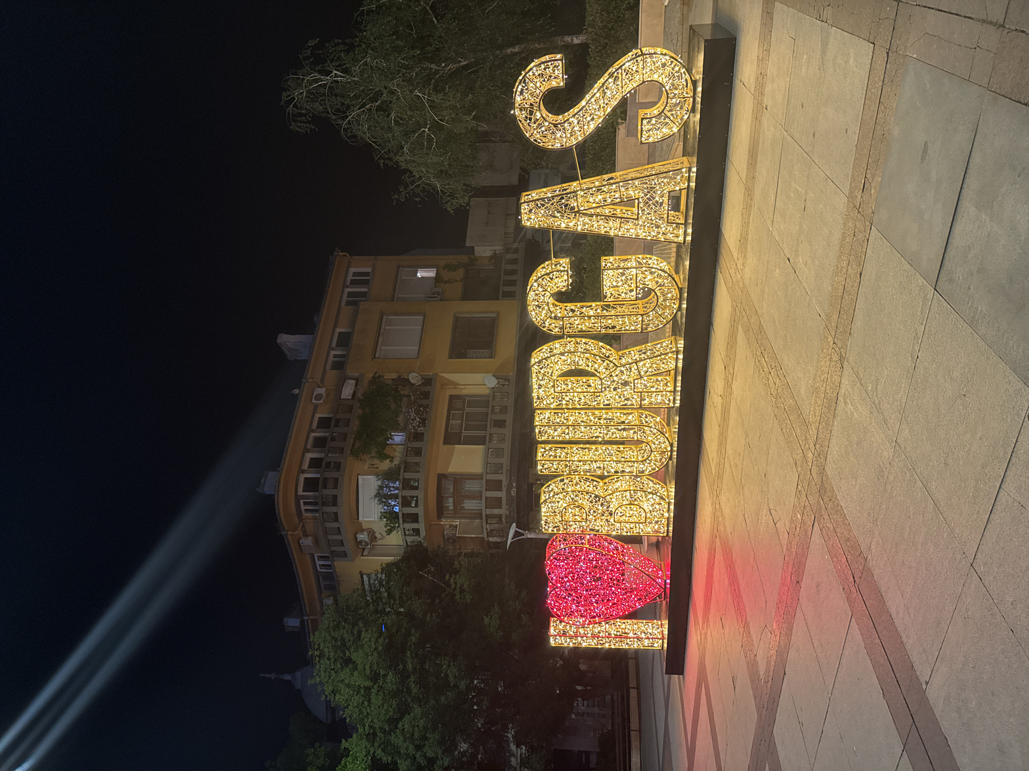 The image shows a night scene in the city of Burgas, Bulgaria, featuring a large illuminated sign that says "I LOVE BURGAS". The sign consists of an illuminated red heart and large, decorative letters spelling out "BURGAs", all adorned with yellow lights. The sign is placed on a paved plaza in front of a yellow building with multiple windows and balconies. Trees are visible on the sides of the building, and in the sky, two bright spotlights are projecting upwards. The ground is paved with stone tiles, with some dark rectangular inlays.