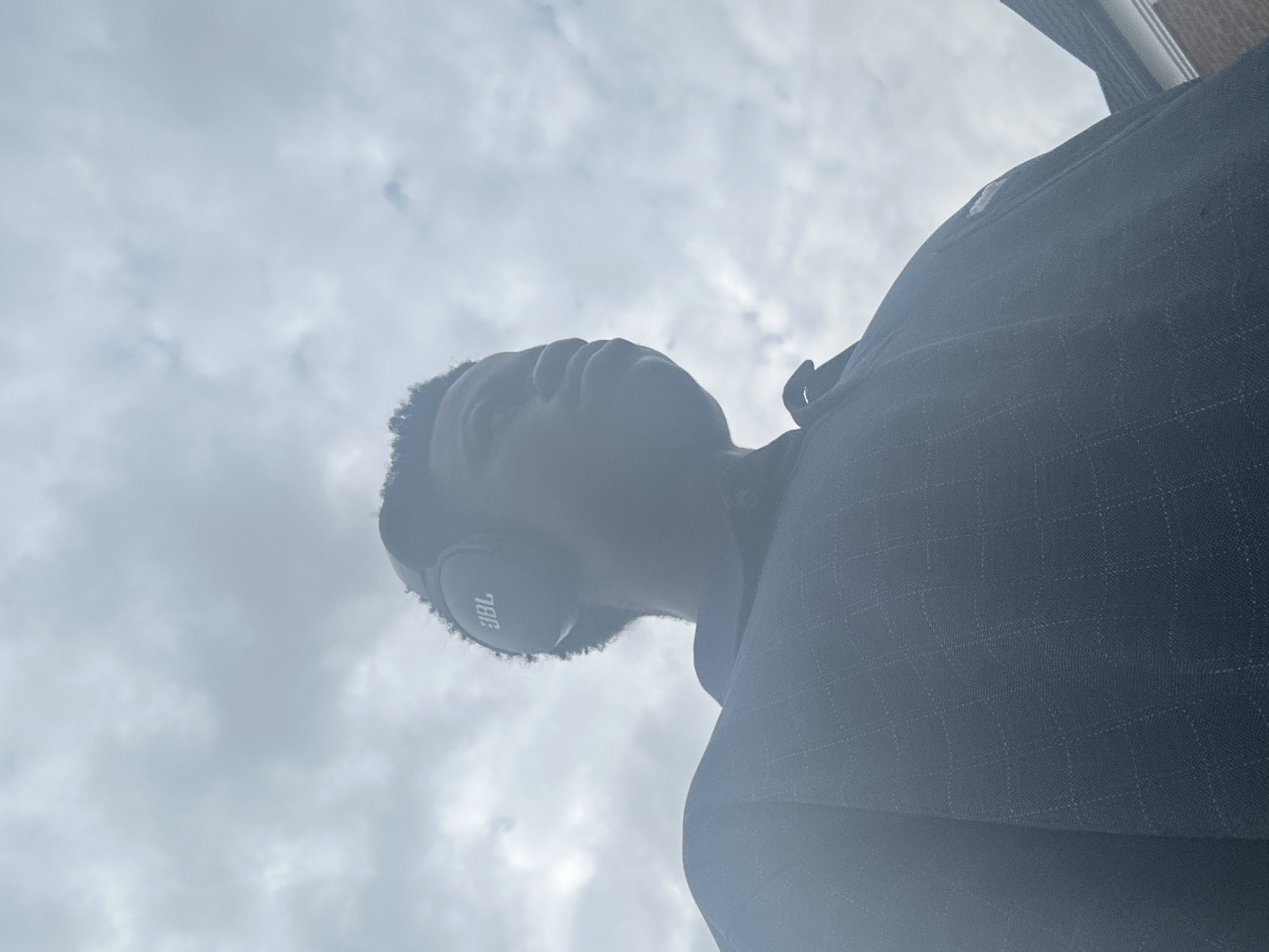 A low-angle, outdoor, waist-up shot shows a young Black person from the chest up, wearing dark headphones with the JBL logo visible, and a dark, collared shirt with a subtle checkered pattern. The person is looking to the right of the frame, with their face in shadow. The background consists of a hazy, overcast sky, with a dark sloped roofline and a portion of a brick wall visible on the right edge of the frame. The image does not provide any specific visual information to determine a location within the city of Badagry, Nigeria.
