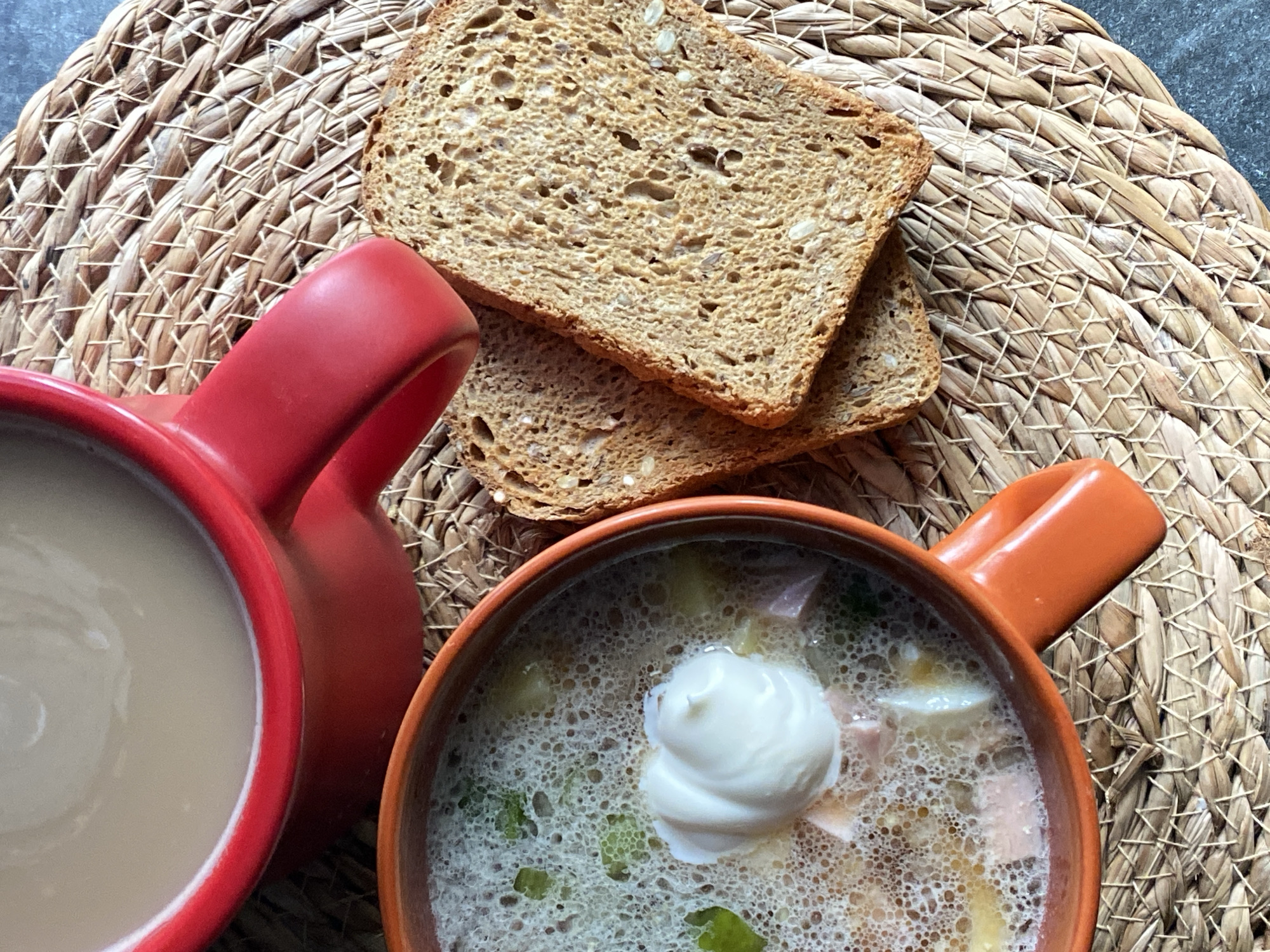 This is a top-down, close-up shot of a simple meal. In the foreground, an orange bowl contains a creamy soup with visible chunks of meat and vegetables, topped with a dollop of white sour cream. To the right, two slices of whole-wheat toast with visible seeds are stacked on a woven placemat. Behind the soup, a red mug holds a light-colored beverage, likely coffee or tea. The entire scene is arranged on a dark, textured surface, possibly a countertop or table. No people are visible, and there are no indications of the specific location in Chaikovsky, Russia, beyond the general setting of a meal.