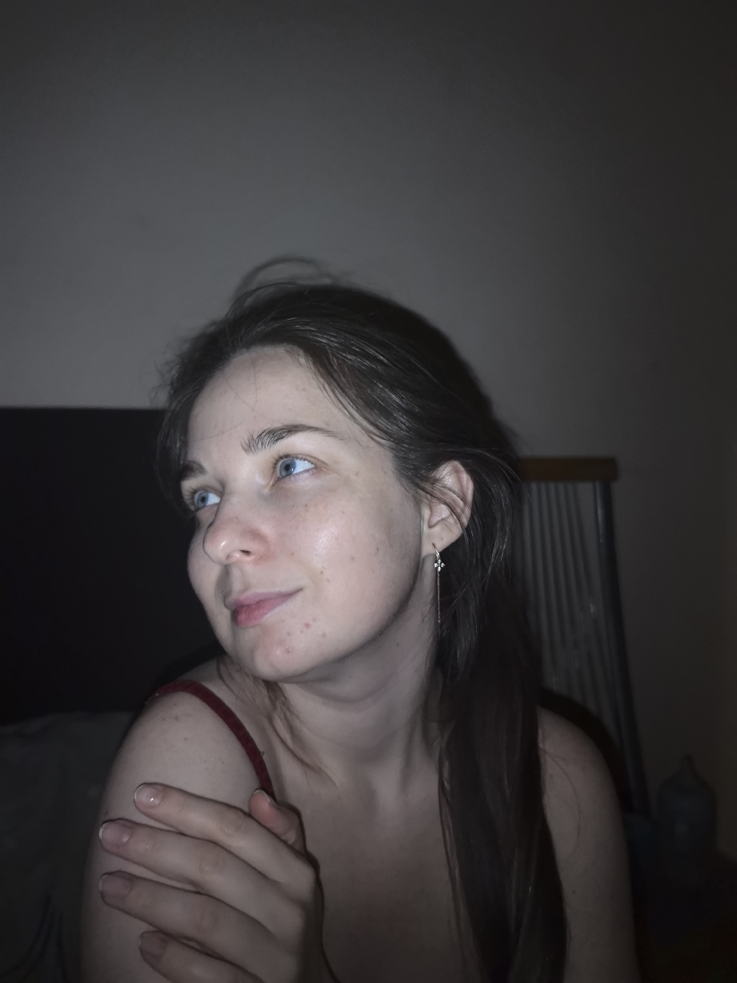 A young woman with fair skin, shoulder-length dark hair, and blue eyes is captured in a dimly lit indoor setting. She is wearing a thin-strapped top and an earring in her left ear. Her left hand is resting on her shoulder. The background is indistinct, appearing to show a wall and possibly furniture. There is no indication in the image that it was taken in Moscow, Russia.