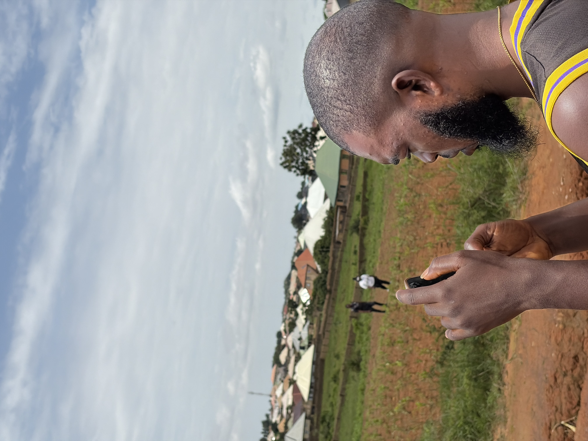 In Rafin Bauna, Nigeria, a man with a close-cropped haircut and a full beard is captured in a profile view, holding a smartphone to take a picture or video. He is wearing a black sleeveless shirt with purple and yellow trim. The background reveals a rural landscape featuring a cultivated field, residential buildings with various roof colors, and a cloudy sky. Two individuals are visible in the middle ground, standing amidst the field. One appears to be wearing a dark shirt and the other a white top. The foreground is dominated by reddish-brown soil and green grass, indicating a dry or arid environment.