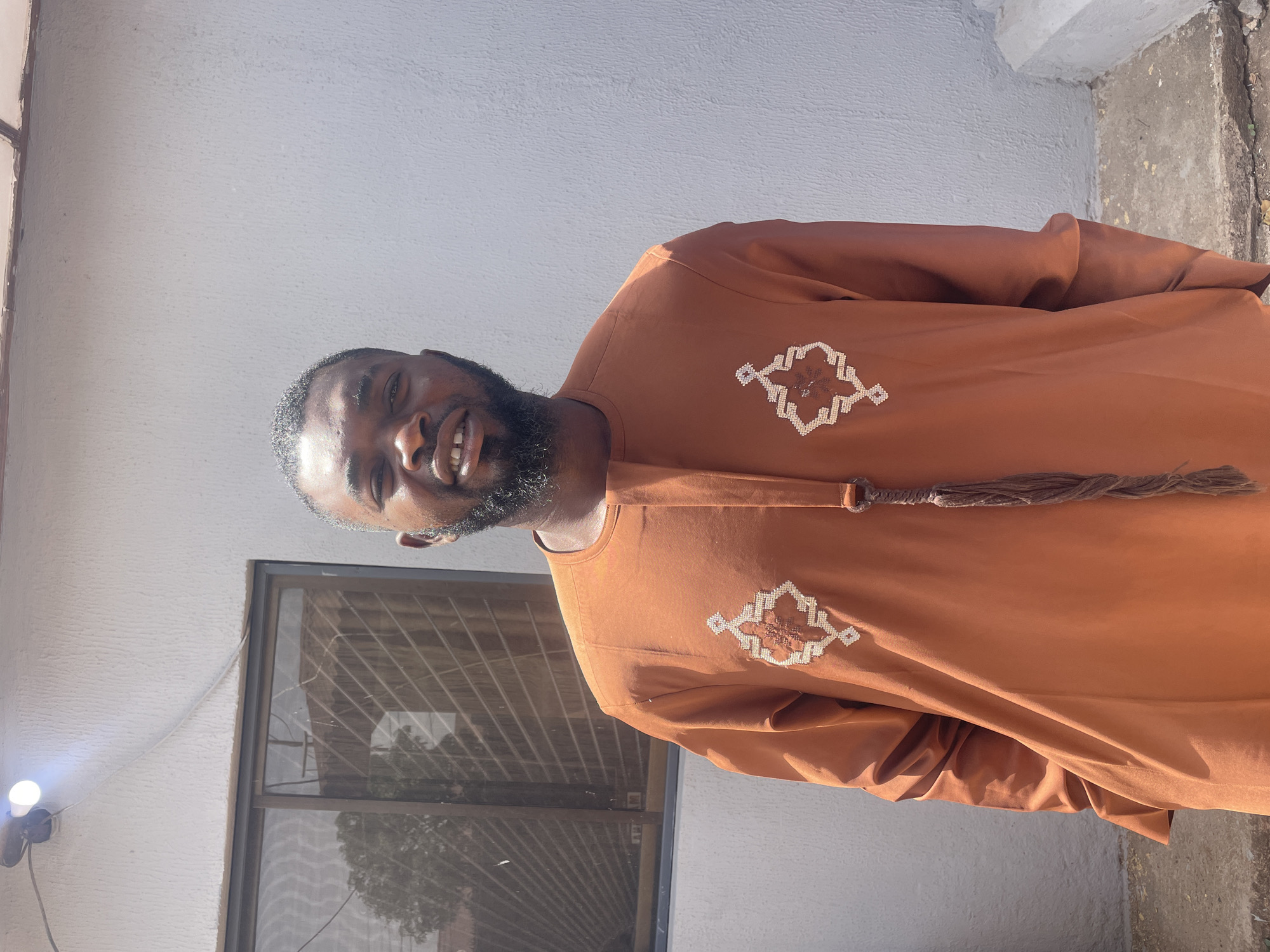 An outdoor, brightly lit, waist-up portrait shows a man smiling. He has dark skin, short dark hair, and a neatly trimmed beard. He is wearing a burnt orange or rust-colored traditional garment with long sleeves and a round neckline. The garment features intricate light-colored embroidery in a geometric pattern on the chest, with two prominent embroidered sections. A decorative, braided tassel in a similar earthy tone hangs from the front of the neckline. The man is smiling widely, showing his teeth, and appears to be in a pleasant mood.
The background consists primarily of a light-colored, textured wall, possibly painted white or off-white. To the man's left, a dark-framed window with a visible security grille or bars is set into the wall. Reflections, possibly of trees, are seen in the window glass. Further to the left, a bright, glowing LED light bulb is attached to the wall with a visible white cable leading from it. A small section of rough concrete ground or a wall base is visible in the lower right corner of the frame. The strong, direct sunlight suggests it is daytime. The scene is set in Jos, Nigeria. No visible text is present in the image.