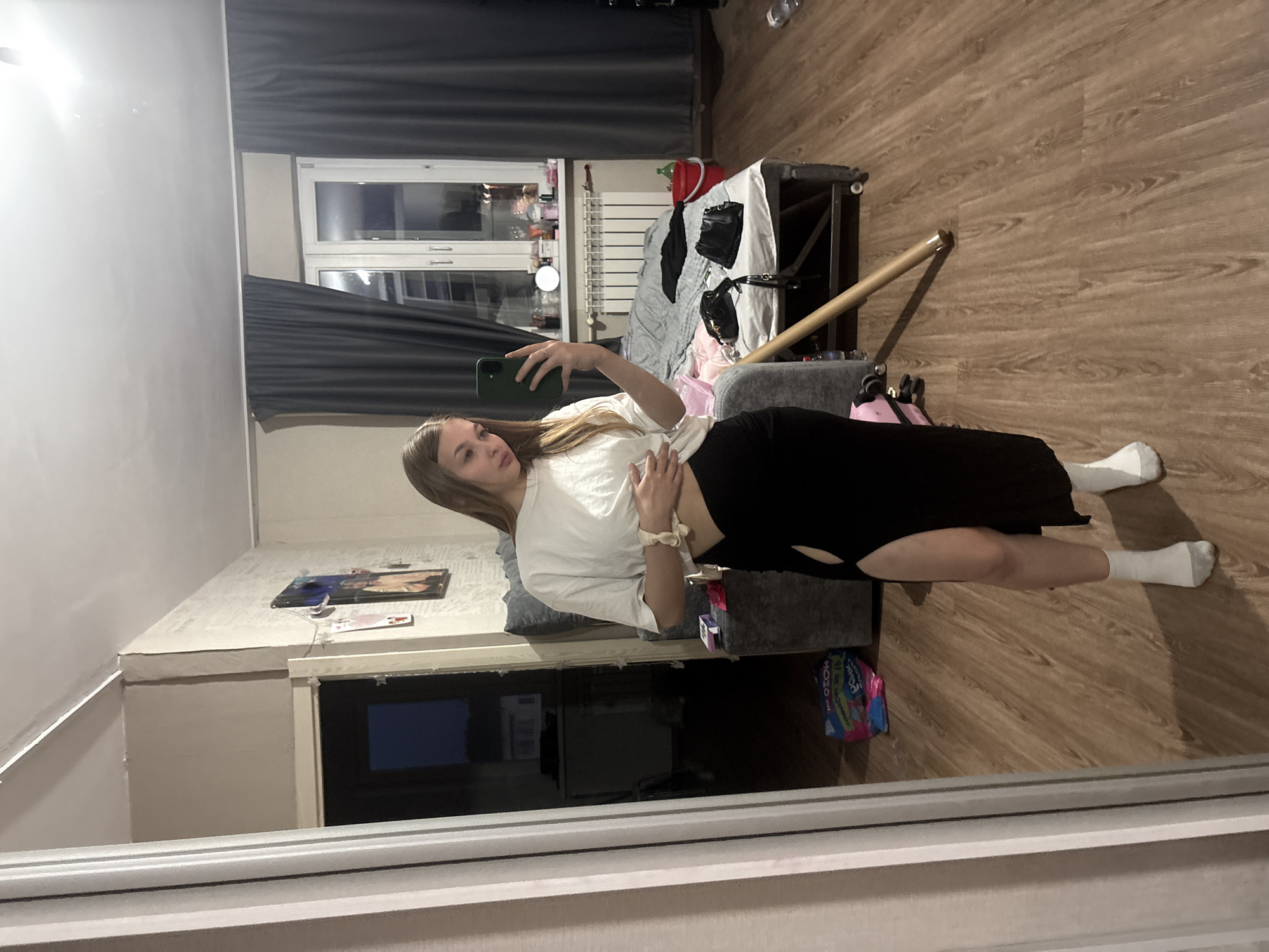 A young woman with blonde hair takes a mirror selfie in a dimly lit bedroom. She is wearing a white t-shirt and a long black skirt with a high slit on the side, white socks, and a scrunchie on her wrist. She is holding a green smartphone in her right hand and resting her left hand on her stomach. The room is cluttered with furniture and personal belongings, including a bed with gray bedding, a dresser with items on top, a wooden floor, and a window with dark gray curtains. There are also some bags and a red bucket visible. The image appears to be taken in an apartment in the city of Chaikovsky, Russia.