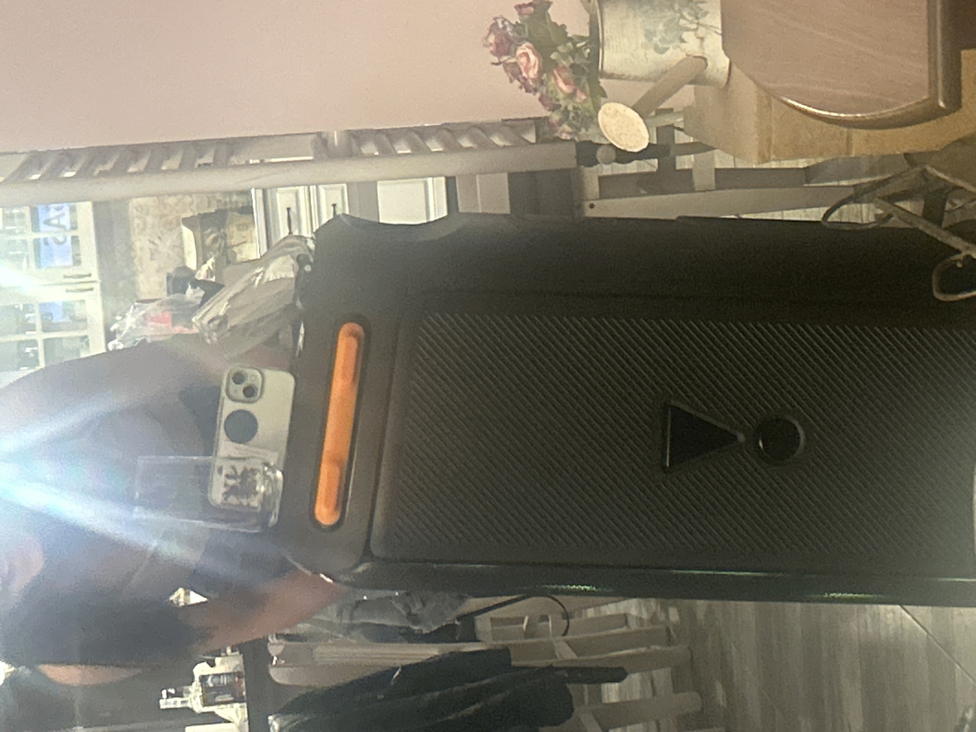 The image shows a black JBL speaker with orange accents. The speaker is positioned on a surface that appears to be a white-painted shelf or rack. A cell phone with a clear case is visible near the speaker, being held by a person. There is also a table, flowers, and a decorative watering can in the background. The location is not apparent from the image.