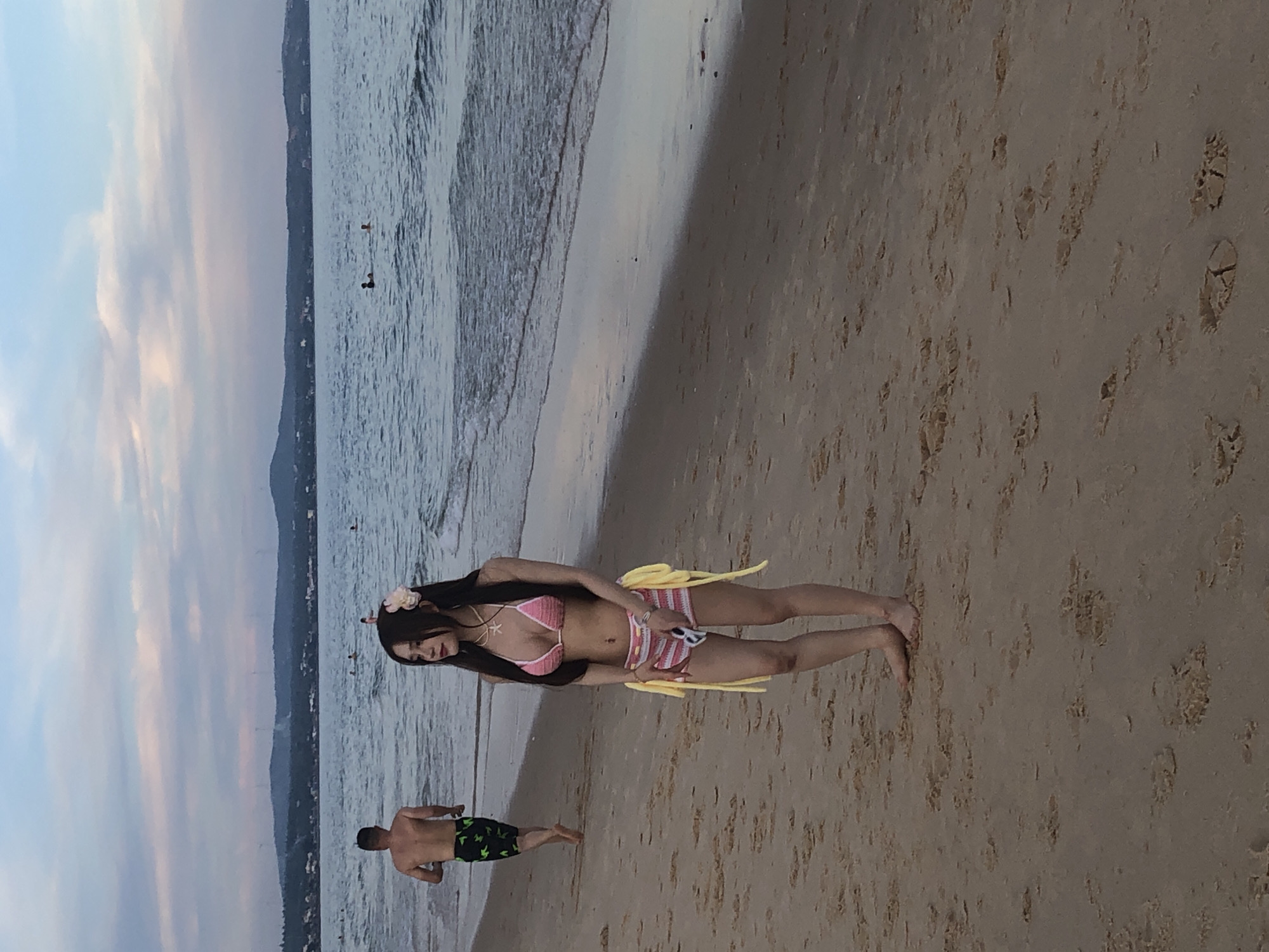 In Phu Thuy Ward, Vietnam, a beach scene unfolds with two individuals. A man, shirtless and wearing green patterned swim trunks, walks away from the camera towards the ocean. To his right and closer to the foreground, a woman stands facing forward, looking slightly to her left. She is wearing a pink crochet bikini with yellow ties and pink and white striped shorts. Her dark hair is partially covered by a pink flower. The beach is sandy, with the gentle waves of the ocean lapping at the shore. In the distance, a hazy coastline with hills can be seen under a cloudy sky.