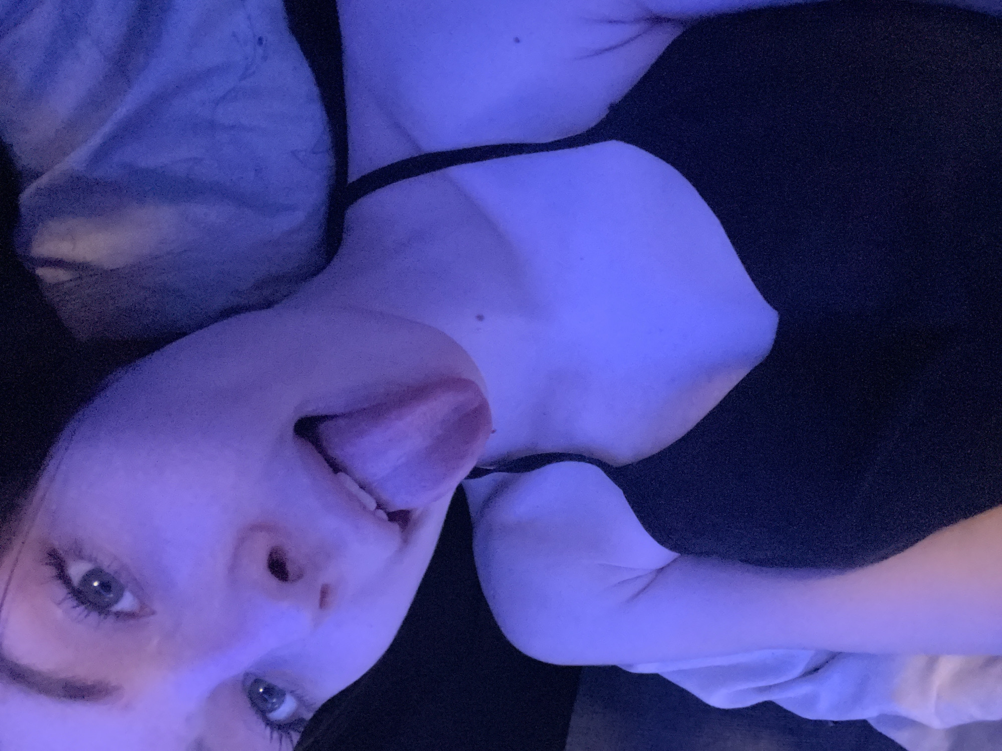The image displays a close-up, angled portrait of a person, likely female, lying down indoors. The scene is bathed in prominent blue and purple artificial lighting, suggesting an evening or nighttime setting.
The subject's head, neck, and upper chest are visible, with the person looking directly into the camera. Their eyes are wide open, and they have a playful or mischievous expression with their tongue sticking out. They appear to have long eyelashes and well-defined eyebrows.
The person is wearing a black top or camisole with thin straps, partially revealing their upper body. Bits of bedding or a pillow are visible on the left side and around the subject, indicating an indoor, bedroom-like environment. The blue light imparts a cool, ethereal tone to the skin. Small moles are discernible on the person's neck and chest. No text is visible in the image. The provided location context of Sinyavino, Russia, is not visually verifiable within the image itself.