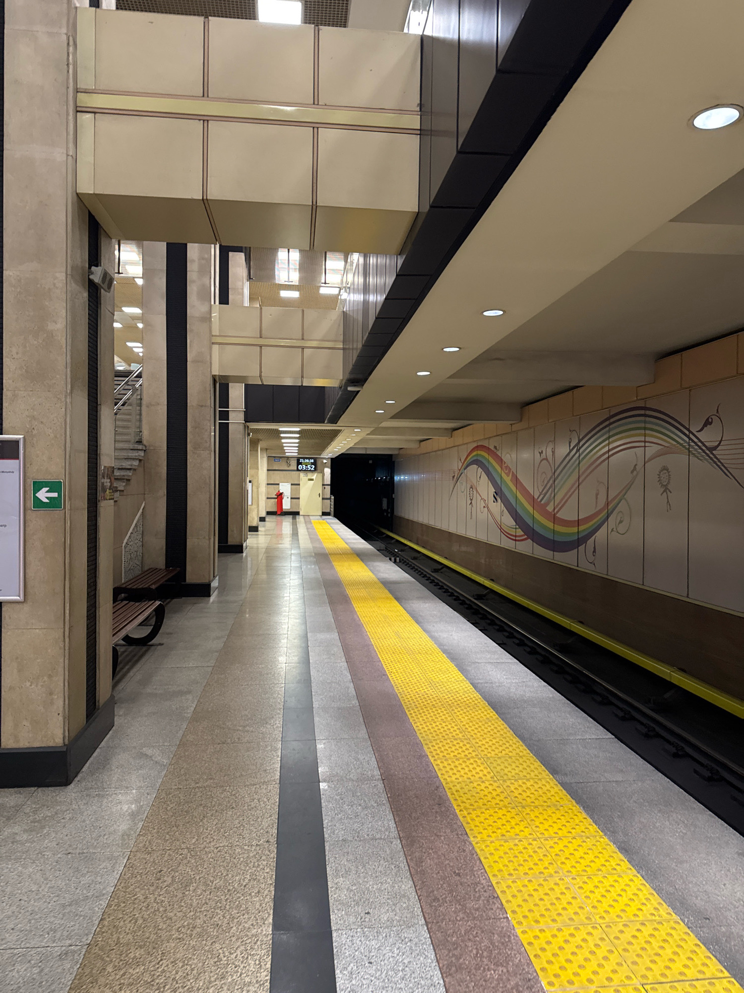This is a vertical, medium-shot, eye-level, full-color photograph of an empty subway platform in Almaty, Kazakhstan. The platform is clean and well-lit, with a textured yellow line running along the edge of the track. To the left, a large stone column supports a multi-leveled ceiling with recessed lighting. A wooden bench is mounted to the column. A green directional arrow sign is visible on a white informational poster mounted to the column. Further down the platform, a digital clock displays the time as 03:52, and a red-clad figure stands near a wall. The wall to the right of the tracks features a mural of a rainbow and abstract designs. The perspective is looking down the length of the platform, creating a sense of depth and emptiness.