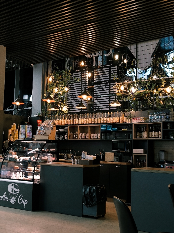 A coffee shop interior is shown. A glass display case showcases pastries. Behind it, a counter stands against shelves filled with bottles, jars, and a microwave. A menu board hangs above, adorned with string lights and greenery. Pendant lights illuminate the scene from a slatted ceiling. A chair is visible in the foreground, suggesting seating for customers. The overall ambiance is warm and inviting, with a modern, industrial aesthetic.