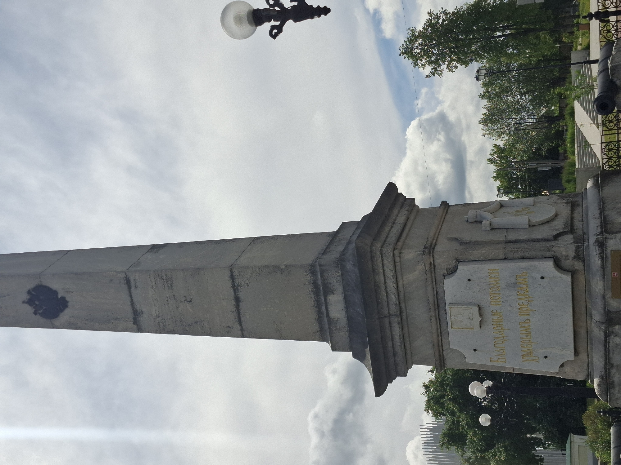 This media file captures a tilted, low-angle view of an obelisk monument with an inscription. The monument itself is made of stone, with a tall, tapering shaft that has a dark, possibly soot or stain, mark on its left side. The base of the obelisk features decorative architectural elements and a prominent stone plaque with golden Cyrillic text. The inscription on the plaque reads: "БЛАГОДАРНЫЕ ПОТОМКИ ХРАНИТЬ ПОДВИГОВЫ".
To the right of the obelisk, a cannon barrel is visible, suggesting a historical or military context. Beyond the monument, a wrought-iron fence encloses a grassy area with trees and lampposts. In the background, modern architecture with a distinctive striped pattern can be seen peeking through the trees. The sky is filled with large, white, puffy clouds, indicating a bright, possibly daytime setting. A decorative streetlamp with a glass globe is also visible at the top of the frame.
The scene appears to be a public space, likely a park or square, commemorating a historical event or individuals. The presence of the obelisk, cannon, and inscription points to a memorial or monument. The weather seems fair, with a mix of sun and clouds. The orientation of the shot, from a low angle looking upwards, emphasizes the grandeur and height of the monument. The location context provided is Kungur, Russia.