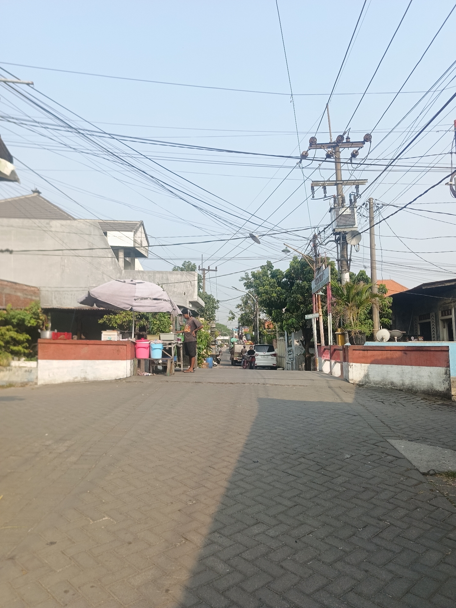 A sunny street scene in Gedangan, Indonesia, features a paved road with interlocking brickwork, shaded by a clear sky. Overhead, a dense network of electrical wires crisscrosses the sky, supported by utility poles. On the left side of the street, a small stall operates under a large grey umbrella, with a person standing beside it, looking towards the right. The stall displays several red and blue plastic buckets. Adjacent to the stall are buildings with grey and white facades, some with tiled roofs. Further down the street, a white car is parked, and two individuals are visible near a bicycle. The right side of the street shows more buildings, some with a red and white painted base, and a palm tree. The overall atmosphere is that of a typical urban street in Indonesia, with evidence of commercial activity and everyday life.