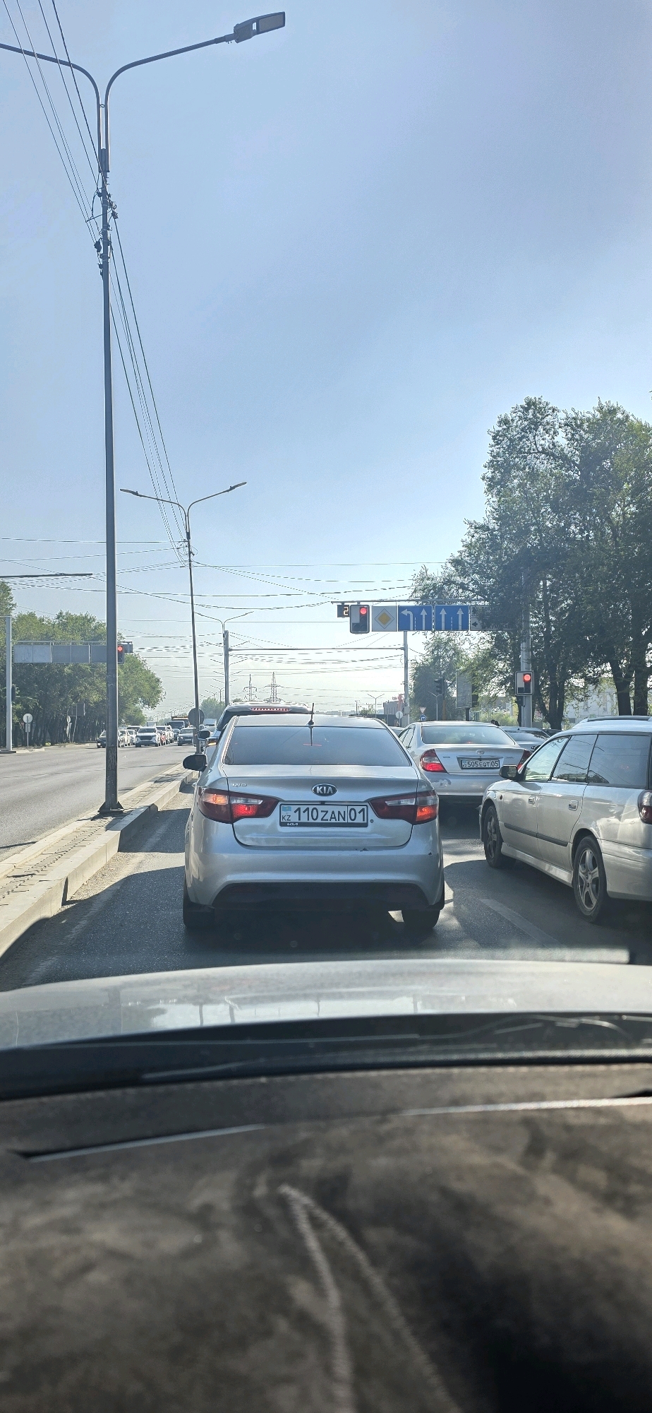 The image captures a view of traffic on a sunny day in Almaty, Kazakhstan. The perspective is from inside a vehicle, looking forward through the windshield, which is dusty. The foreground is dominated by the dark interior of the vehicle.
In the middle ground, a silver Kia sedan is prominent, displaying a license plate that reads "KZ 110 ZAN 01". To the right of the Kia, a silver Subaru Outback is visible. Other cars are present in the traffic jam, indicating a busy road.
The background features a city street with multiple lanes of traffic. Traffic lights and street lamps are visible. Overhead power lines crisscross the sky, a common sight in urban areas. Tall green trees line the sides of the road, suggesting an urban park or residential area. The sky is clear and blue, with a few wispy clouds. The overall scene depicts a typical urban traffic situation in Almaty.