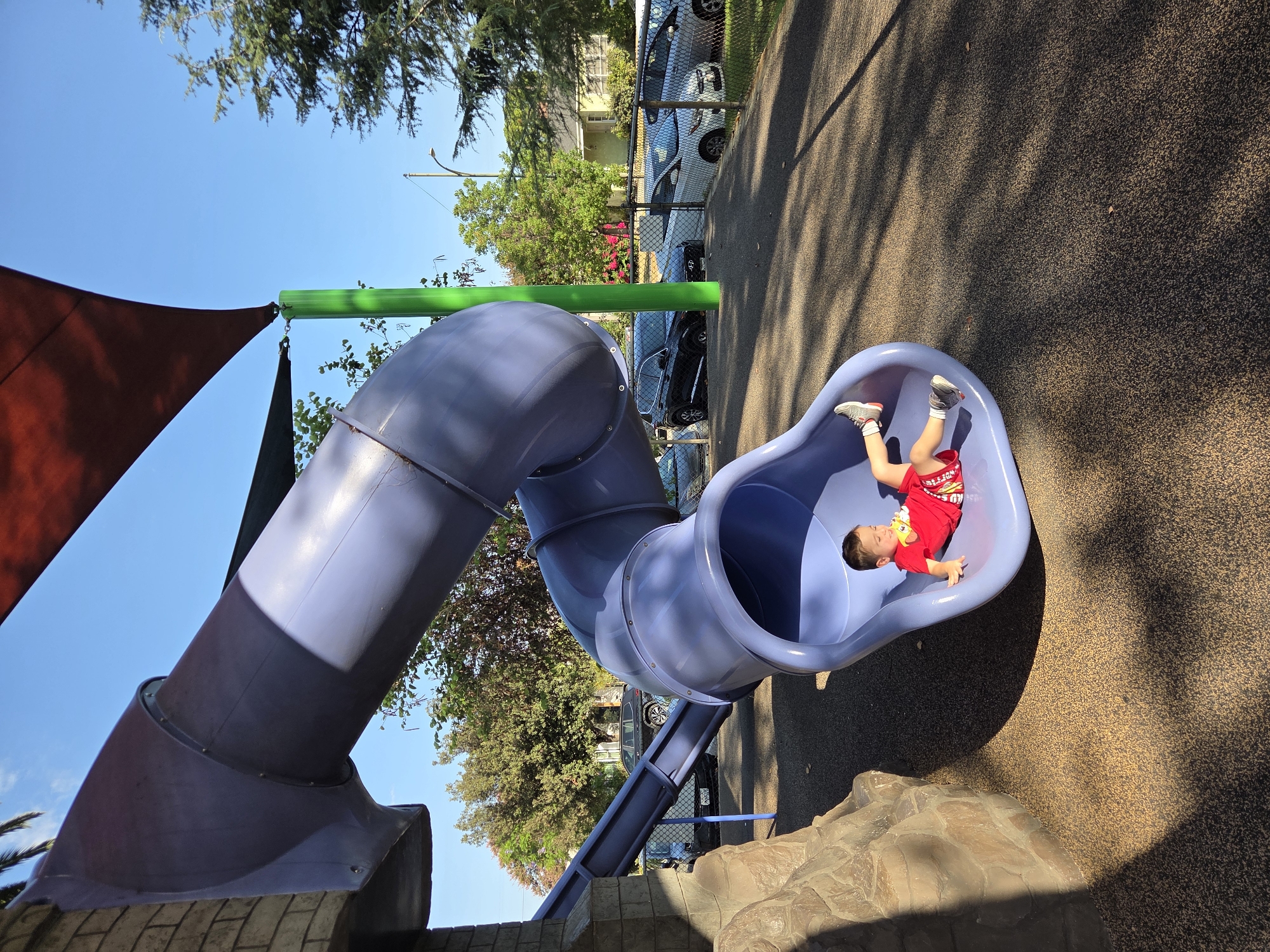 A young boy, wearing a red t-shirt and red shorts with a white pattern, is lying down on his back on a purple plastic slide. His feet are pointed upwards, and his head is turned towards the left side of the image. The slide is part of a playground structure, which includes a faux rock climbing wall on the left. The playground surface is dark brown with visible tan specks. In the background, a chain-link fence encloses parked cars and residential houses. Trees with green and brown foliage are visible, and a clear blue sky dominates the upper portion of the image. A large, dark red shade structure is visible over the playground. The lighting suggests it is daytime, with shadows cast across the playground surface. The scene appears to be located in Glendale, United States of America.