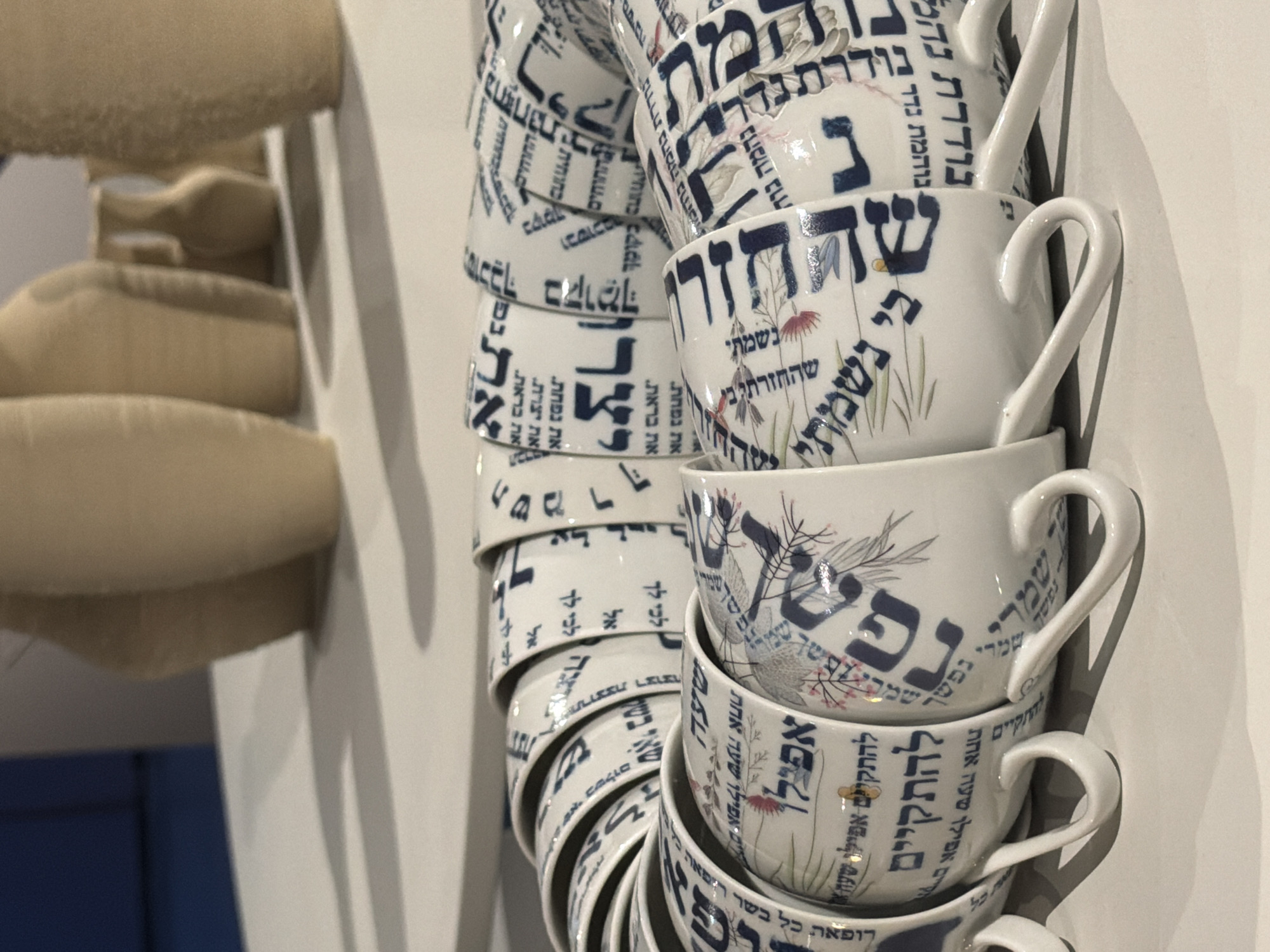 The image shows a stack of decorative tea cups with Hebrew script and floral designs, likely found in a shop in Tel-Aviv, Israel. The cups, predominantly white, feature blue Hebrew lettering alongside delicate floral illustrations. The script appears to be poetic or religious in nature, commonly found in Jewish cultural artifacts. The cups are stacked unevenly against a light gray wall, casting soft shadows. In the blurred background to the left, there is a chair with a soft, beige upholstery.