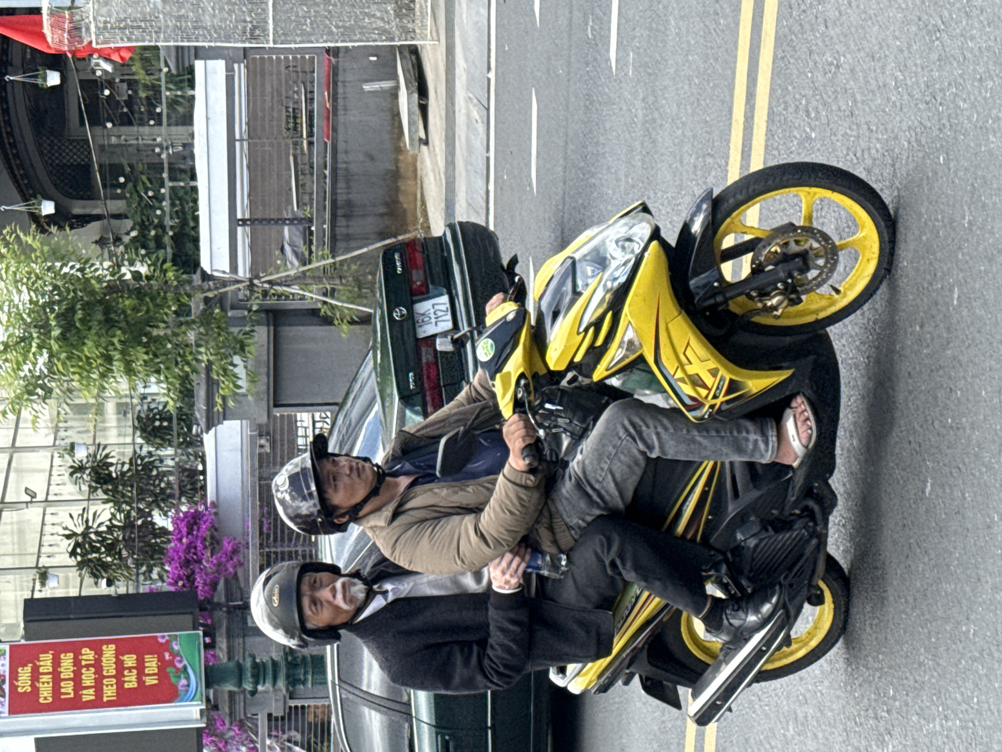 Two men ride a yellow and black scooter on a paved street in Da Lat, Vietnam. The man in the front, driving, wears a black helmet and a brown jacket. The man behind him, the passenger, wears a grey helmet and a dark jacket over a white shirt. He holds a bottle of water. A green car is parked on the side of the road behind them. To the left of the scooter is a sign with Vietnamese text and images of flowers. The street is marked with yellow lines.