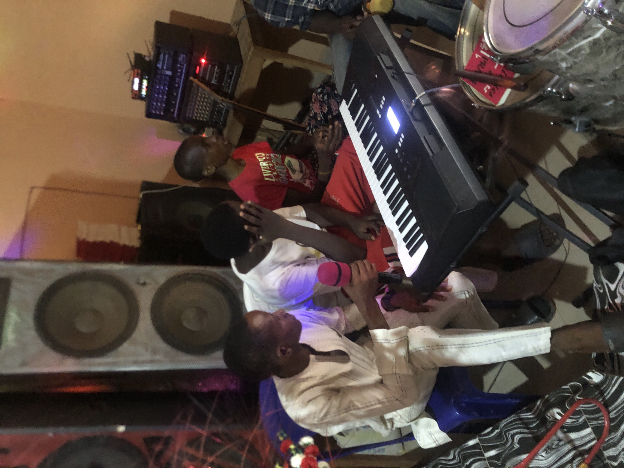 A slightly tilted high-angle shot depicts a musical ensemble in a room, location Nkwelle Ezunaka, Nigeria. A man dressed in a white outfit is seated on a blue plastic chair and holds a pink microphone, possibly singing. Another individual in a white shirt stands next to him, shielding his face with his hand. Two additional people are visible in the background, one wearing a red shirt with a graphic print and the other wearing a red shirt with writing on it. The ensemble is positioned around a black keyboard on a stand. The room is equipped with sound and musical equipment, including speakers with large circular drivers, black rack-mounted units, and drums. The flooring appears to be made of light-colored tiles or concrete.