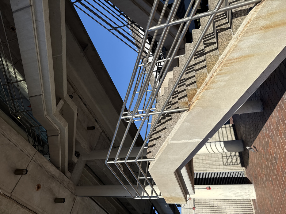An architectural shot, looking upward, featuring concrete structures, metal railings, and a glimpse of blue sky. Concrete supports and beams form sharp angles against the sky, while a metal staircase with railings cuts diagonally across the frame. The composition emphasizes the interplay of lines and shadows, and the contrasting textures of concrete and metal. The shot's orientation creates a sense of disorientation and emphasizes the height of the structures.