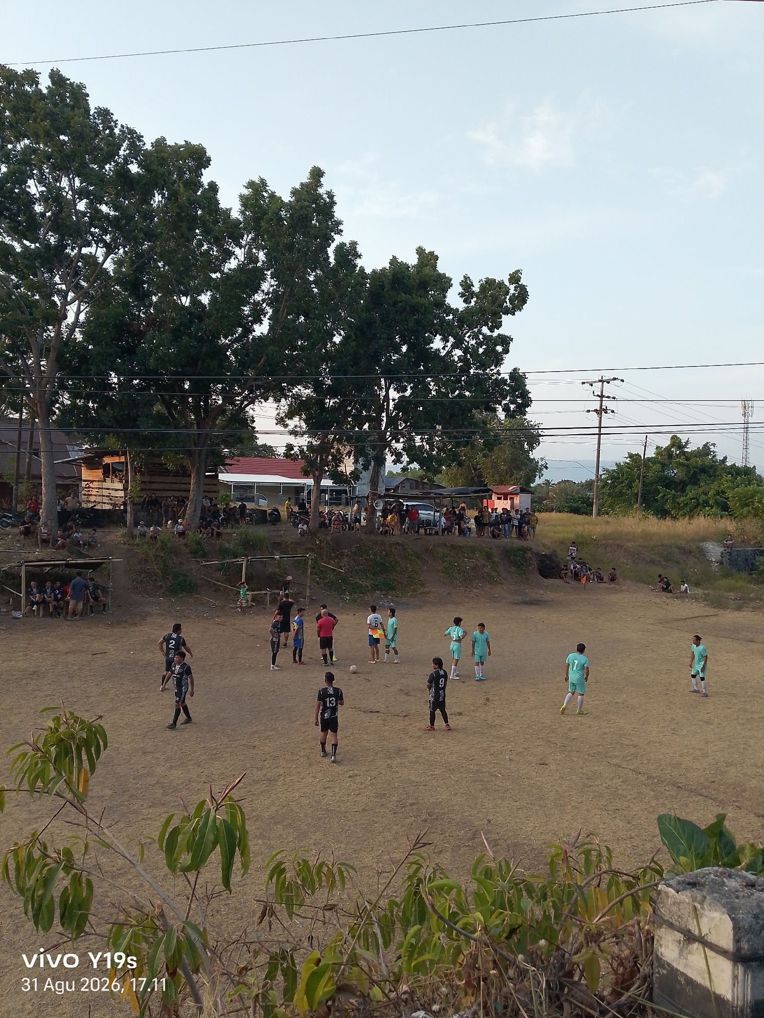In the city of Ranoyapo, Indonesia, a group of young men are playing a soccer game on a dirt field. The field is surrounded by trees and houses, and there are spectators watching the game from the sidelines. The game is in full swing, with players running and kicking the ball. The sky is a clear blue, and the sun is shining. A watermark in the bottom left corner reads "vivo Y19s 31 Agu 2026, 17.11".