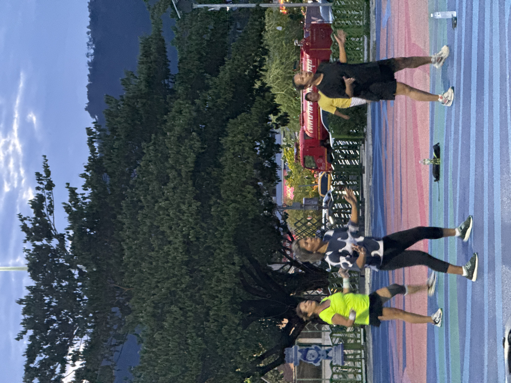 The image depicts an outdoor fitness class in a public area, likely a park or promenade, in Ko Samui District, Thailand, during twilight hours. Three women are actively participating in what appears to be an aerobics or dance routine on a brightly colored, striped surface.
In the foreground, a woman on the right is wearing a black t-shirt and shorts, with athletic shoes, and her arms are extended as if demonstrating a move. To her left, another woman in a black and white patterned shirt and black leggings is also engaged in the activity, her body angled and arms in motion. Further to the left, a third woman, wearing a neon green t-shirt and a black skirt with knee support, is also participating in the exercise.
The background reveals a large, dense tree, and beyond that, hints of urban elements such as a red fire truck and other vehicles are visible, suggesting a relatively populated area. The sky is a dusky blue with some clouds, indicating the transition from day to night. Two water bottles are placed on the ground near the participants, and a small black bag is also visible. The overall scene conveys a sense of communal activity and recreation in a tropical evening setting.