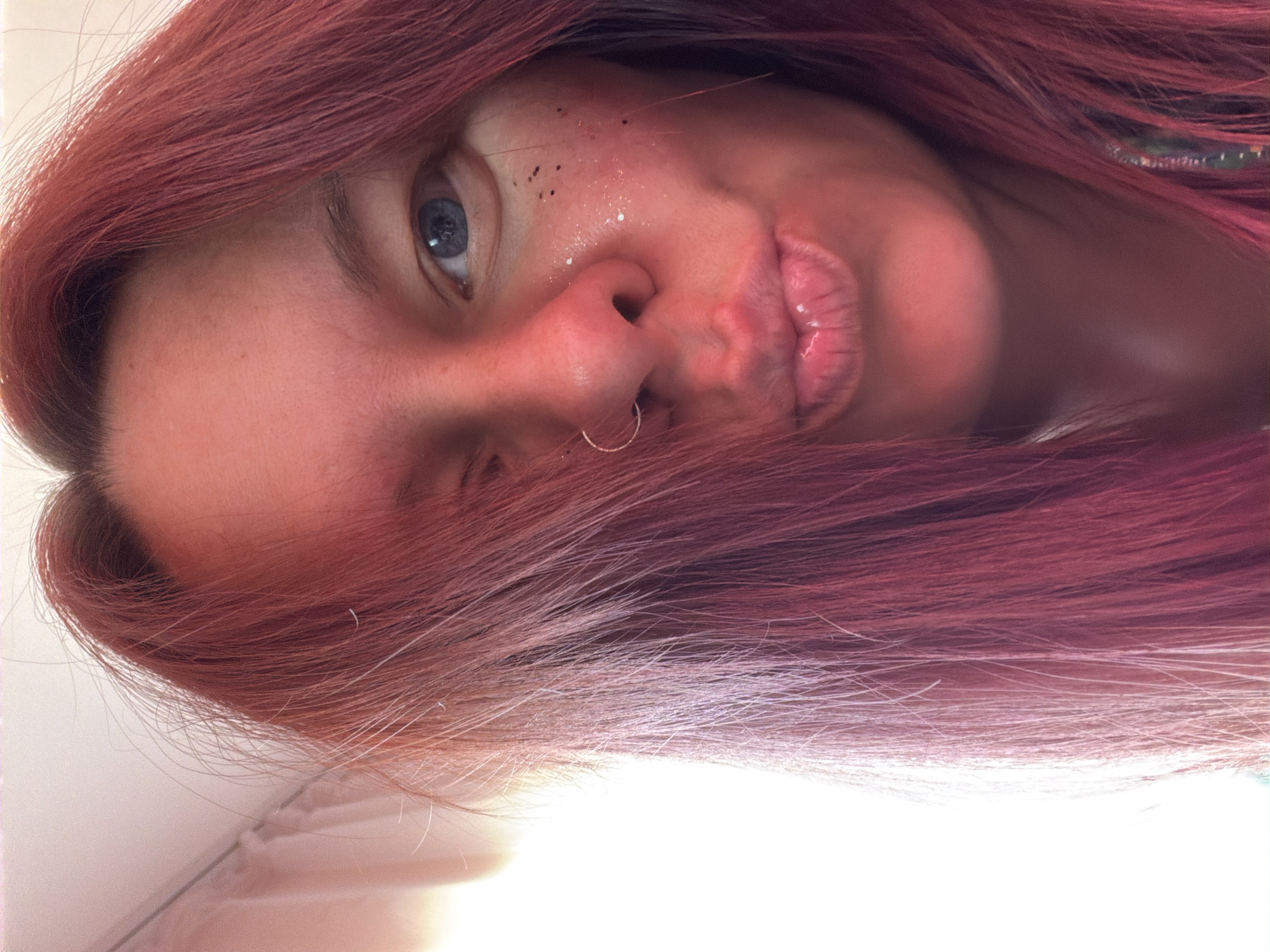 A close-up, angled portrait of a person with red-tinted hair. The person has fair skin, light blue eyes, and wears a nose ring. They have small, dark spots near their eye, possibly freckles, and some shimmering cosmetic glitter on their face. The background is out of focus and bright, possibly white or light-colored. The photo is likely taken indoors, but there are no identifiable features to place it in Chaikovsky, Russia, or any other specific location.