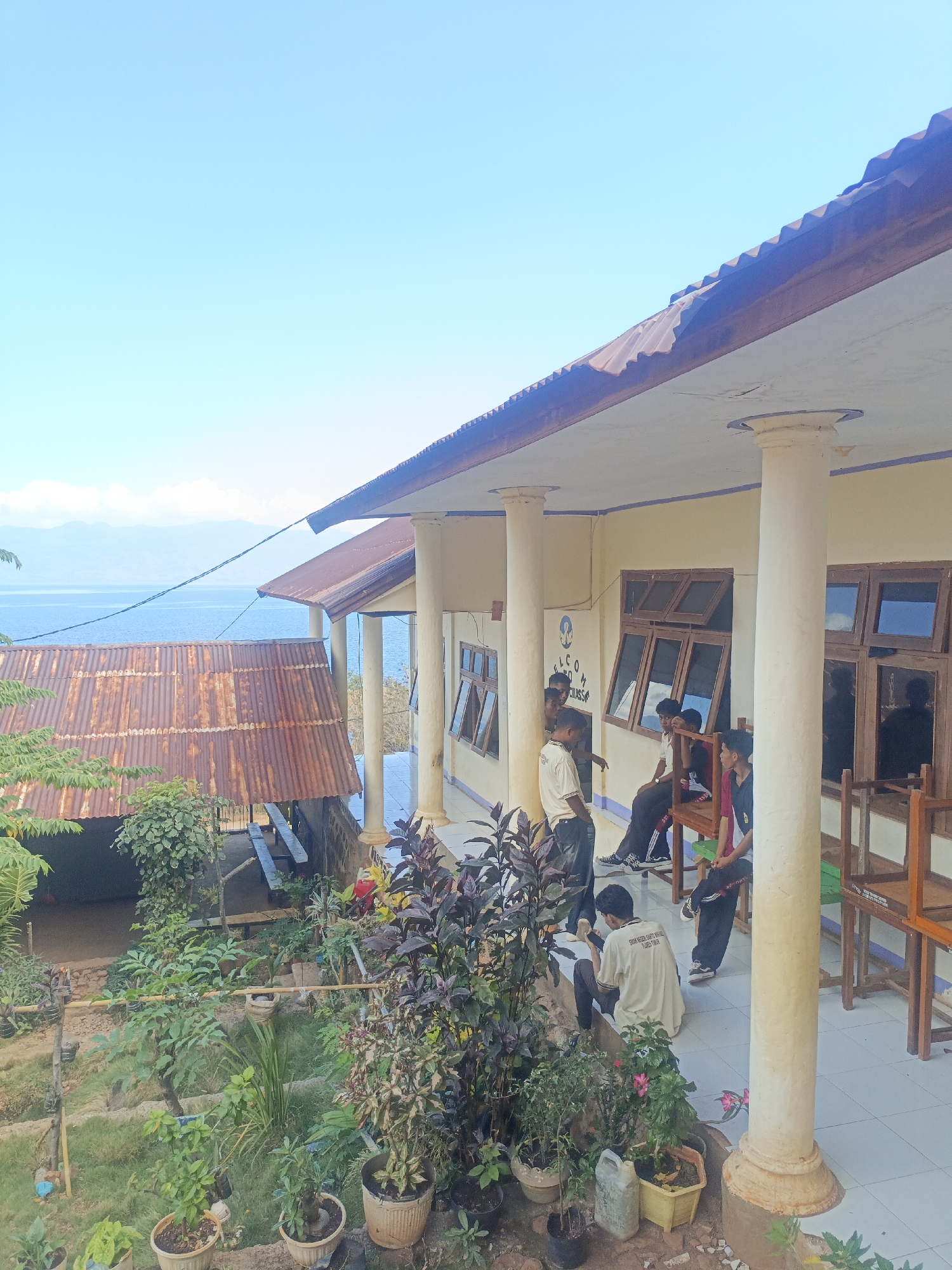 The image depicts a scene near a building with a veranda overlooking a body of water, likely the ocean, with mountains in the distance. The sky is clear and blue. On the veranda, several young males are gathered, some seated, others standing, engaged in conversation or looking at a mobile phone. The building appears to be a school or administrative facility, indicated by a sign reading "SELCOM CLASS" and a logo. In the foreground, there is a lush garden with various potted plants and greenery. A structure with a rusty corrugated metal roof is visible to the left, adjacent to the main building. The overall impression is of a casual gathering in a tropical or subtropical environment, possibly during the daytime. The location is specified as Balaweling I, Indonesia.