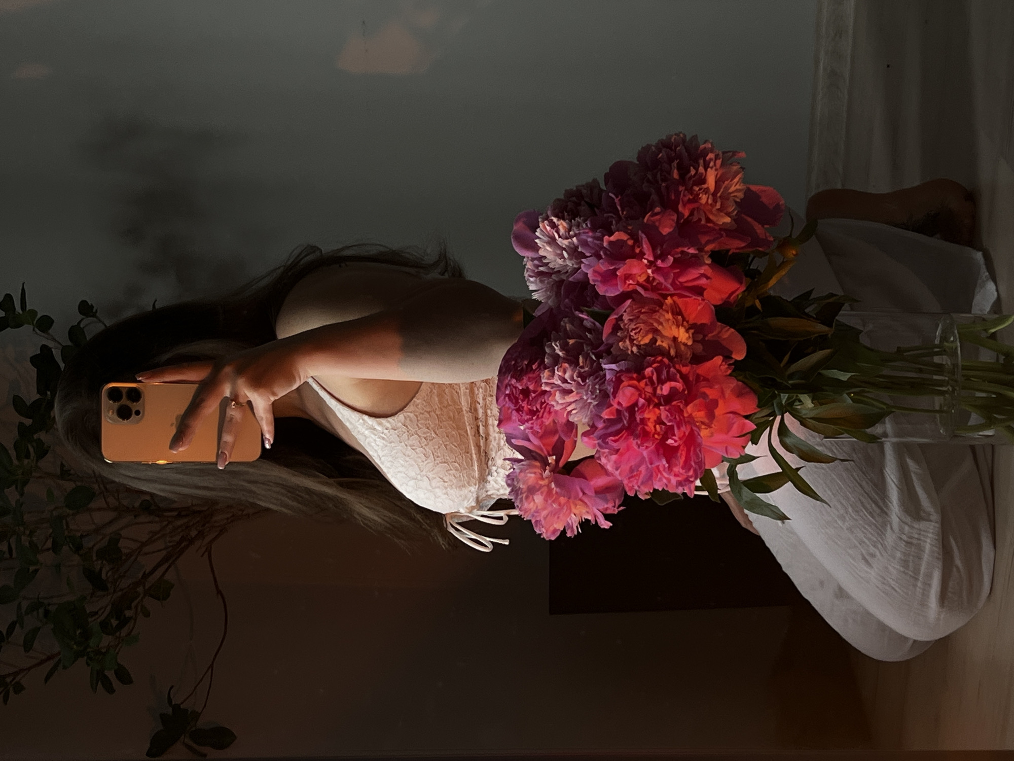 In a dimly lit room with warm, artificial light casting shadows, a person is captured in a mirror selfie. The individual is kneeling on the floor, holding a bouquet of vibrant pink and purple peonies in a glass vase. They are wearing a white lace top and loose white pants. The person is holding a golden smartphone to take the photo, obscuring their face. A plant with dark green leaves is visible in the background, to the left of the frame. The overall setting suggests a personal and intimate moment, possibly in a home environment in Ternopil, Ukraine.
