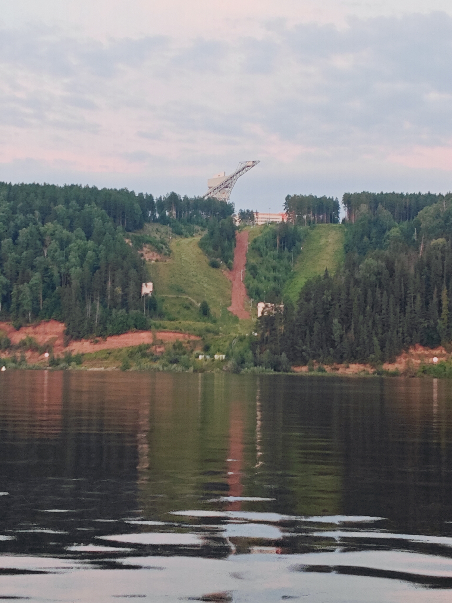 The image depicts a wide river in the foreground, with its surface reflecting the sky and the forested hills on the opposite bank. The sky is overcast with pastel hues of pink and grey. On the distant bank, a dense forest covers the slopes of hills. A ski jump structure, with its characteristic ramp and support tower, is prominently visible atop one of the hills. A dirt path or track runs down the side of the hill, suggesting a ski slope or training area. There are some buildings at the base of the ski jump structure, and smaller structures are scattered along the shoreline. The overall impression is of a natural landscape with man-made recreational facilities. The location is identified as Noviy, Russia.