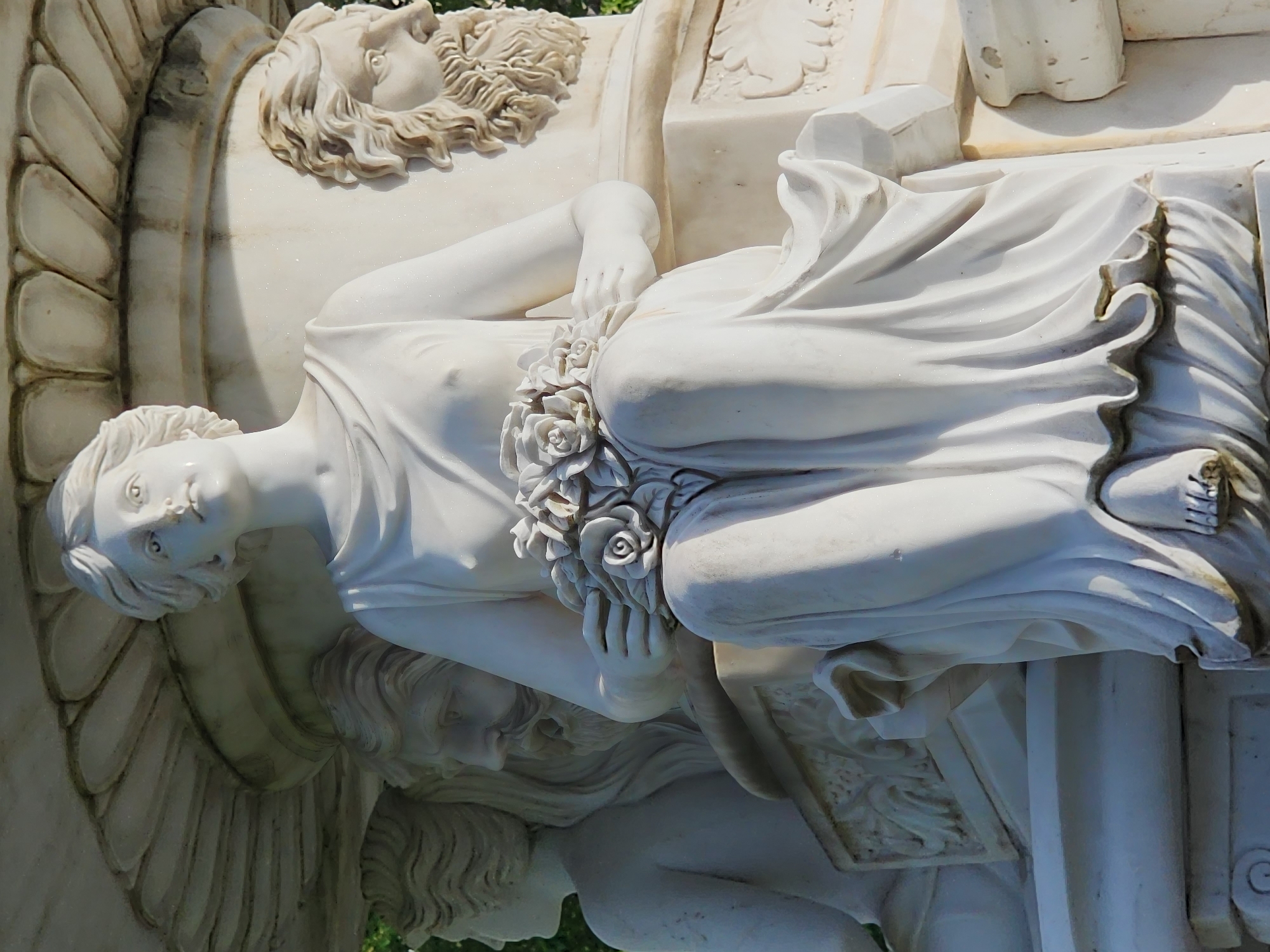 This vertical photograph showcases a section of a monumental white marble sculpture, likely situated outdoors in a park or garden in Sochi, Russia. The sculpture is rendered in a classical style, illuminated by bright daylight, casting distinct shadows and highlights.
The central figure is a gracefully reclining female, possibly a nymph or goddess, depicted with serene features and her gaze directed gently upwards and to the left. Her hair is softly waved, and she is draped in flowing robes that accentuate her form. Her left arm rests across her torso, cradling a small bouquet of intricately carved roses. Her right leg is bent, with her foot visible.
Below this main figure, a partially visible male figure in marble, with curly hair and a beard, appears to recline beneath or support the female. Above and to the right of the central figure, another male head, also bearded and with curly hair, is carved in relief, looking upwards.
The craftsmanship is notable, with realistic drapery folds and delicate floral details. The white marble has a subtle sparkle under the sunlight, suggesting a polished finish, though some minor discoloration is visible in crevices, indicative of age or outdoor exposure. Classical architectural elements, such as fluted or shell-like patterns, form part of the background structure. The strong natural lighting defines the three-dimensional forms, confirming it is daytime. The sculpture conveys a timeless sense of beauty and tranquility. No visible text is present.
