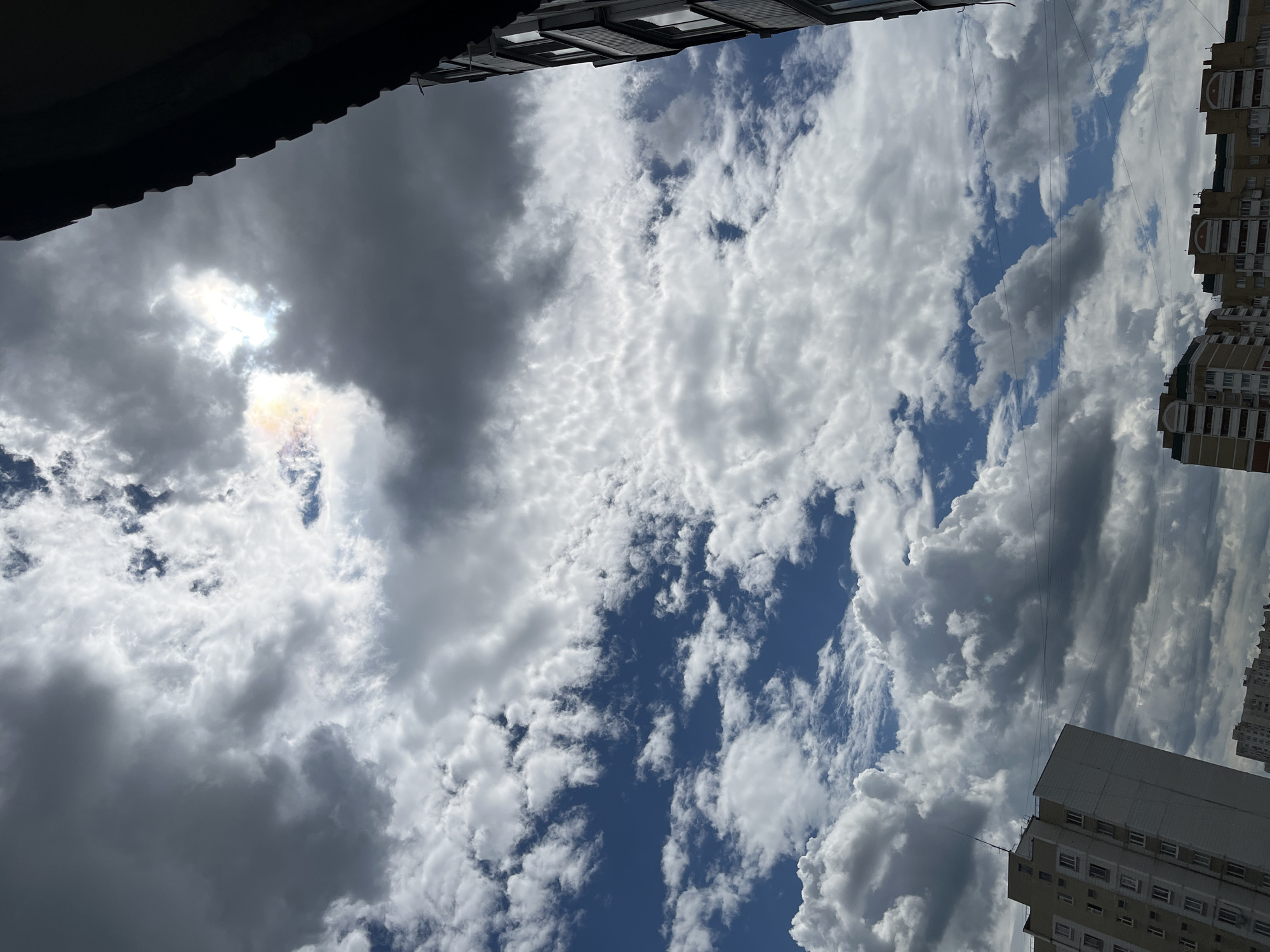 The image shows a view of buildings and the sky in Moscow, Russia. The sky is filled with a mix of white and gray clouds, with patches of blue visible. Sunlight is breaking through the clouds, creating a bright area with a slight rainbow effect. In the foreground, the dark silhouette of a building's edge is visible on the right side of the frame. On the left side, the corner of a light-colored apartment building with multiple windows can be seen. In the distance, other apartment buildings with colorful facades are visible. The overall scene suggests an urban environment with a dynamic sky.