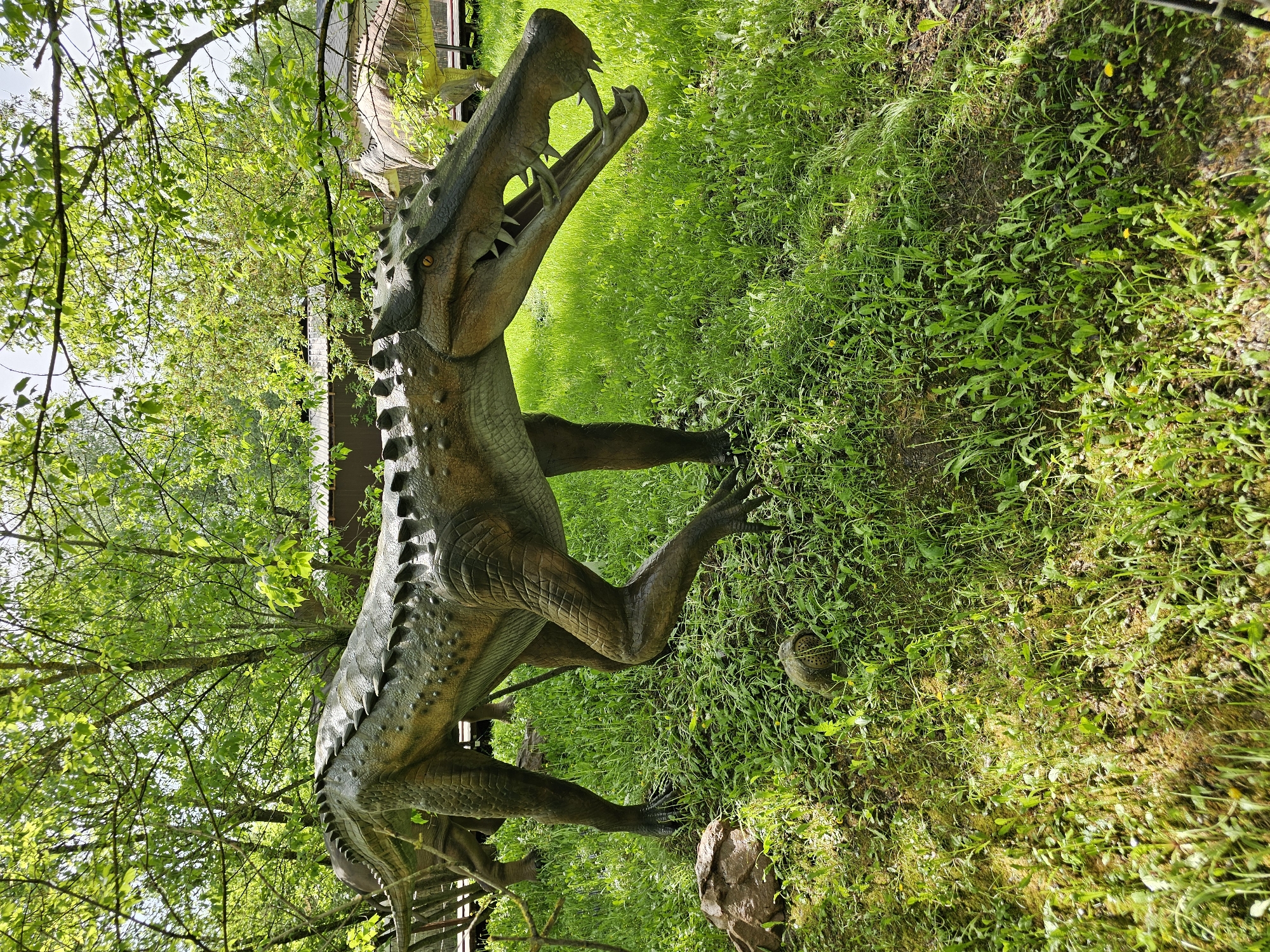 The image depicts a statue of a large prehistoric reptile, possibly a Postosuchus, standing in a grassy area. The statue is detailed with armored plates along its back and sharp teeth visible in its open mouth. The surrounding environment consists of lush green grass and trees with dense foliage. The statue is located in Khimki, Russia, based on contextual information (not visible in the image).