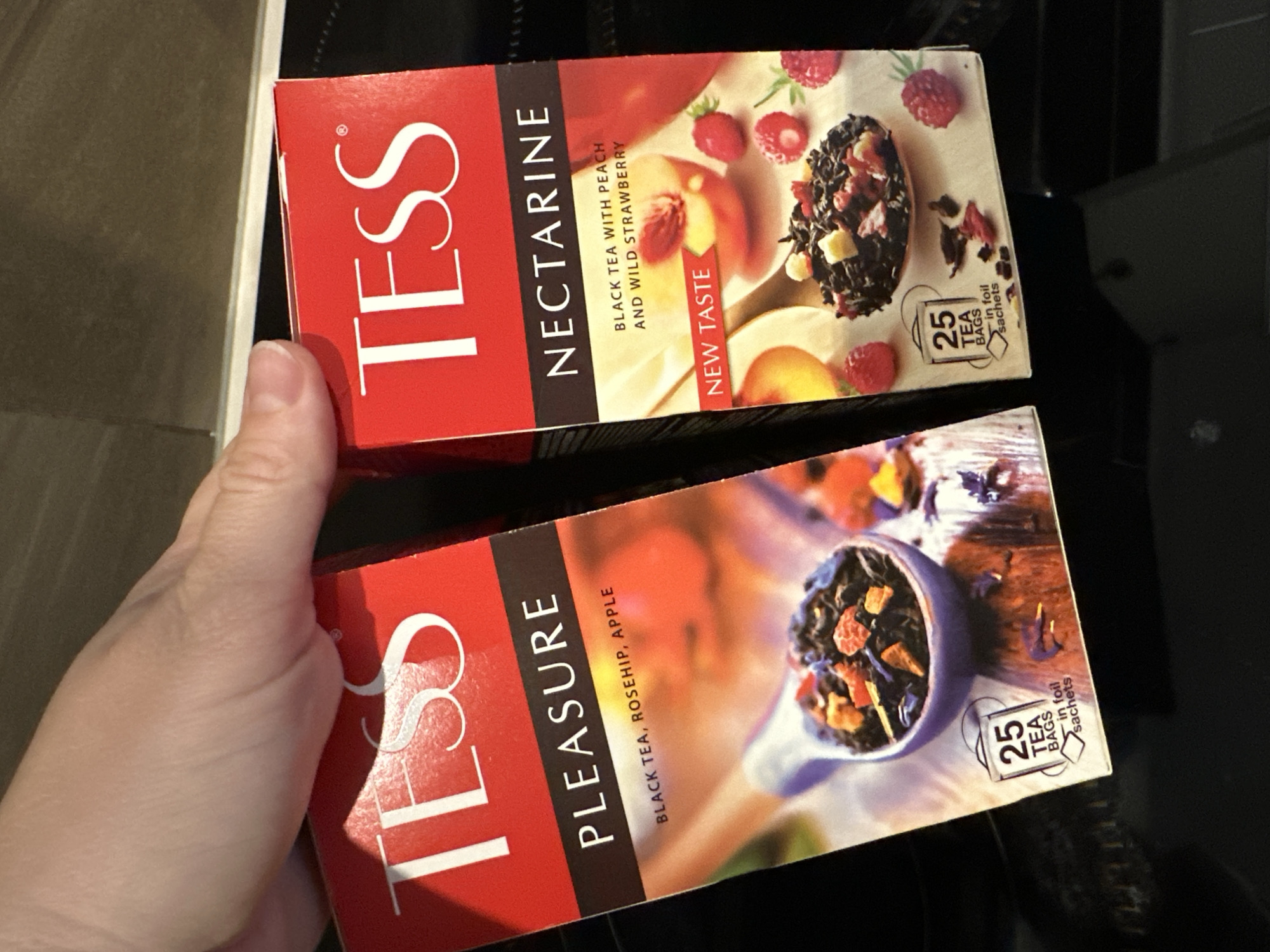 Two boxes of Tess tea are being held by a hand. The box on the left is labeled "TESS PLEASURE BLACK TEA, ROSEHIP, APPLE" and features an image of tea leaves in a spoon. The box on the right is labeled "TESS NECTARINE BLACK TEA WITH PEACH AND WILD STRAWBERRY" and features images of nectarines and raspberries. Both boxes indicate "25 TEA BAGS in foil sachets". The background is dark and out of focus, suggesting an indoor setting.
