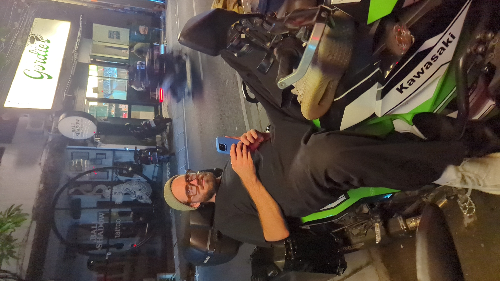 A man with a beard and glasses is sitting on a green and black Kawasaki motorcycle at night. He is wearing a green beanie, a dark t-shirt, and dark pants. He is holding a blue smartphone in his right hand. His left leg is extended forward, resting on the motorcycle. The motorcycle has a black storage box on the back. In the background, there are several shops, including "Gordie's" and "Bali Shadow Tattoo". Motor scooters are parked and moving on the street. The scene appears to be a street in North Kuta, Indonesia.