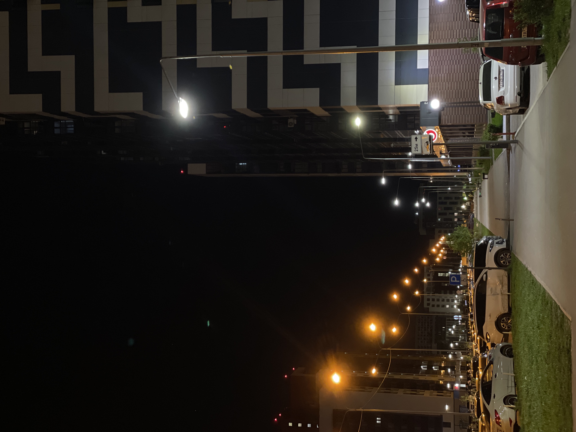 This nighttime photograph depicts a street scene in Kazan, Russia. The street is lined with modern buildings, characterized by geometric patterns of alternating light and dark panels. Streetlights illuminate the scene, casting an orange glow on the road and sidewalk. Parked cars line the right side of the street, adjacent to a strip of grass. Traffic signs and street markings are visible. The sky is dark, contrasting sharply with the illuminated street scene.